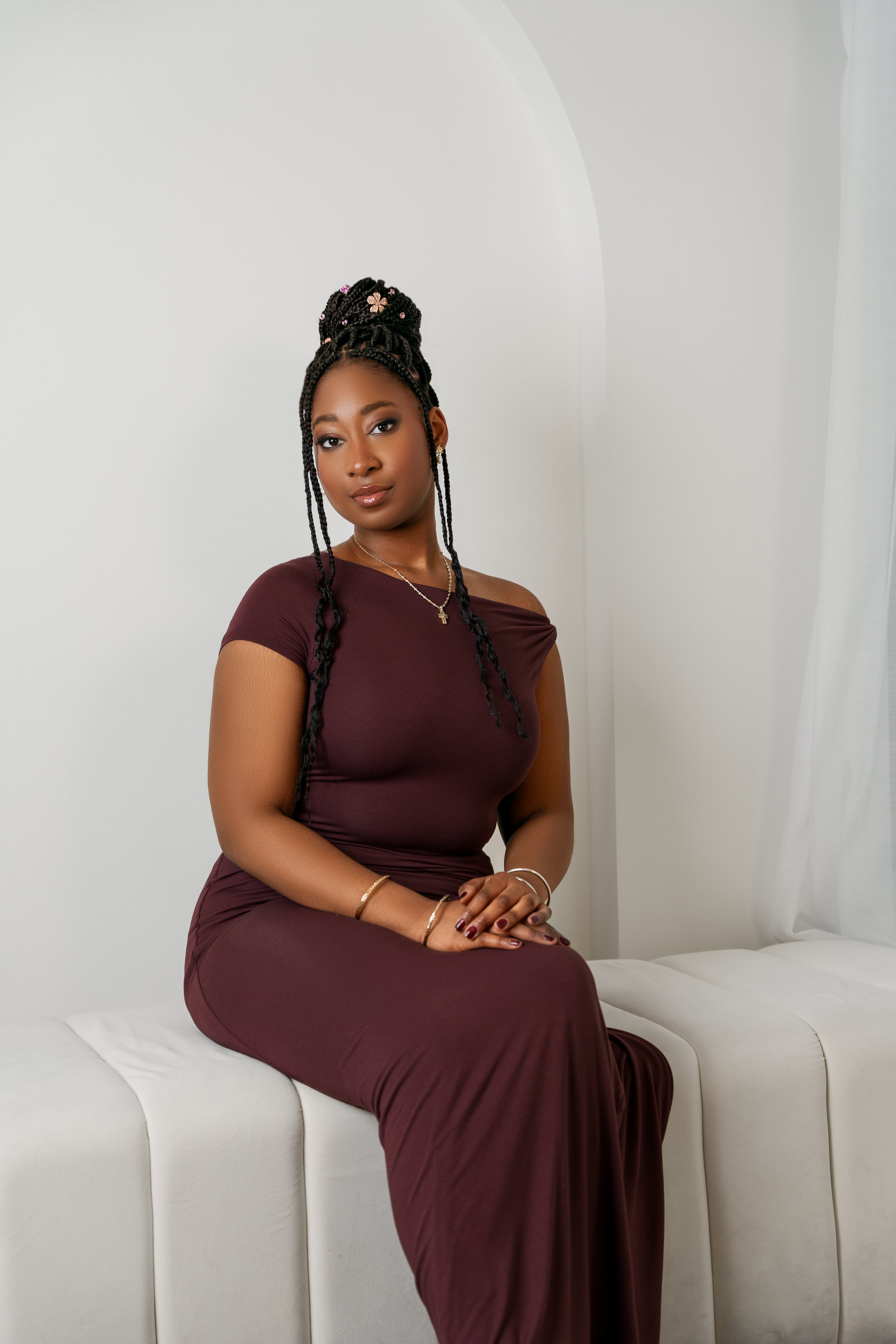 A woman sitting on a white cushioned bench against a plain white wall, wearing a maroon dress, with styled braids decorated with beads and flowers, and accessorized with jewelry.