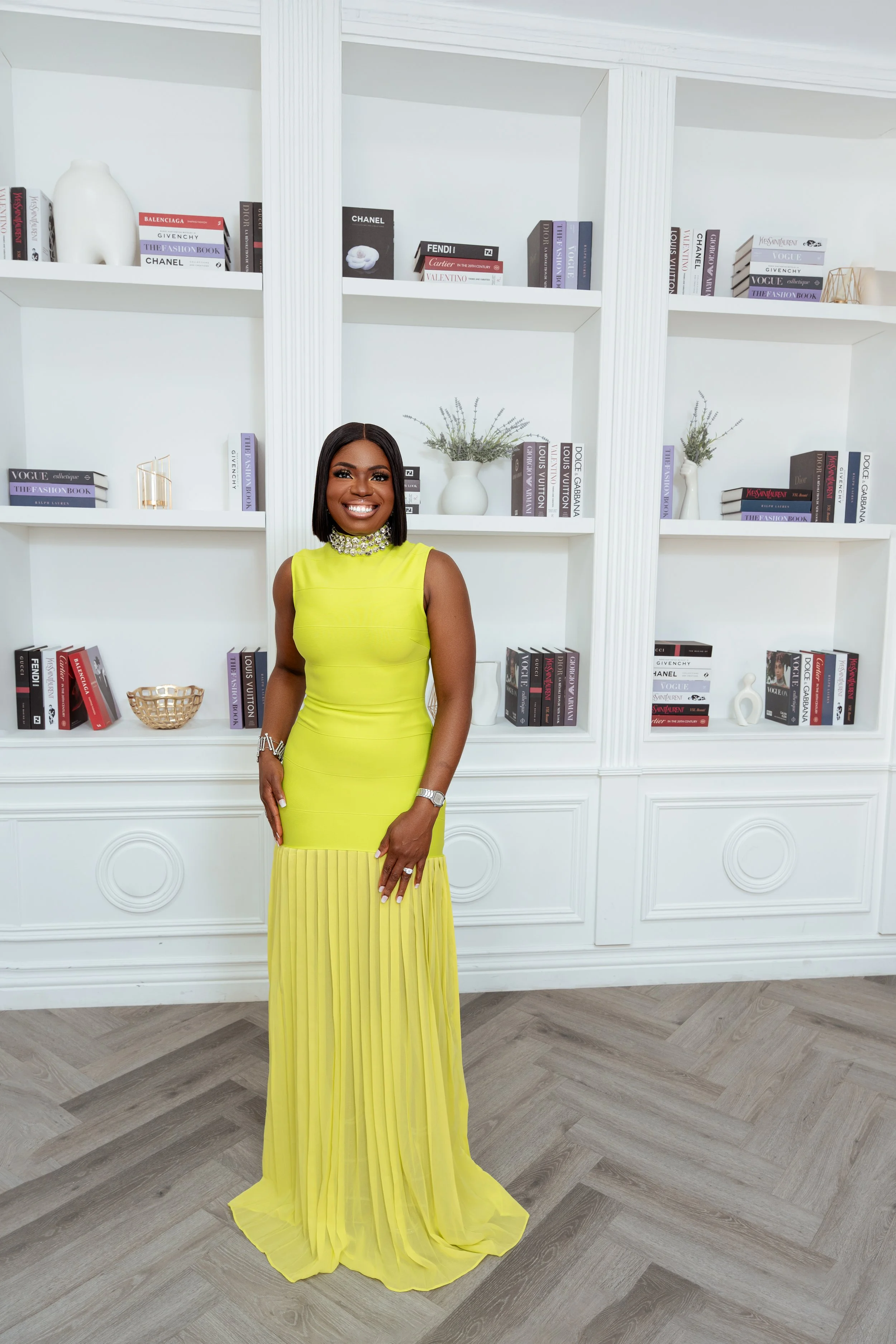 A woman smiling and wearing a bright yellow, sleeveless, pleated gown with a bejeweled collar, standing in front of a white bookshelf filled with fashion and designer books, white vases with lavender, and decorative objects. The floor has a herringbo