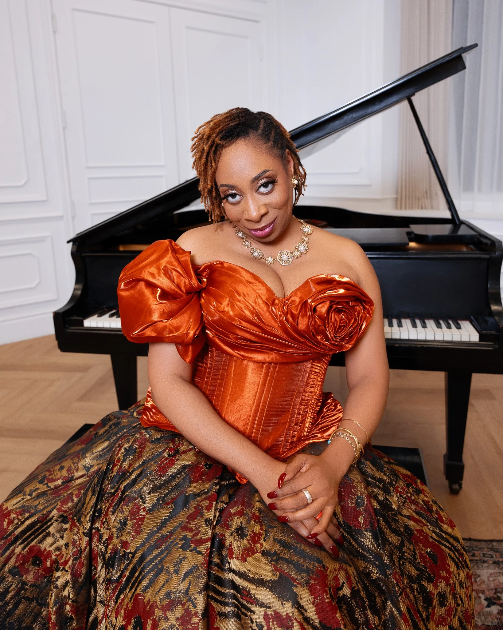 A woman wearing an orange off-the-shoulder gown with large puffed sleeves, sitting in front of a grand piano with a white wall background.