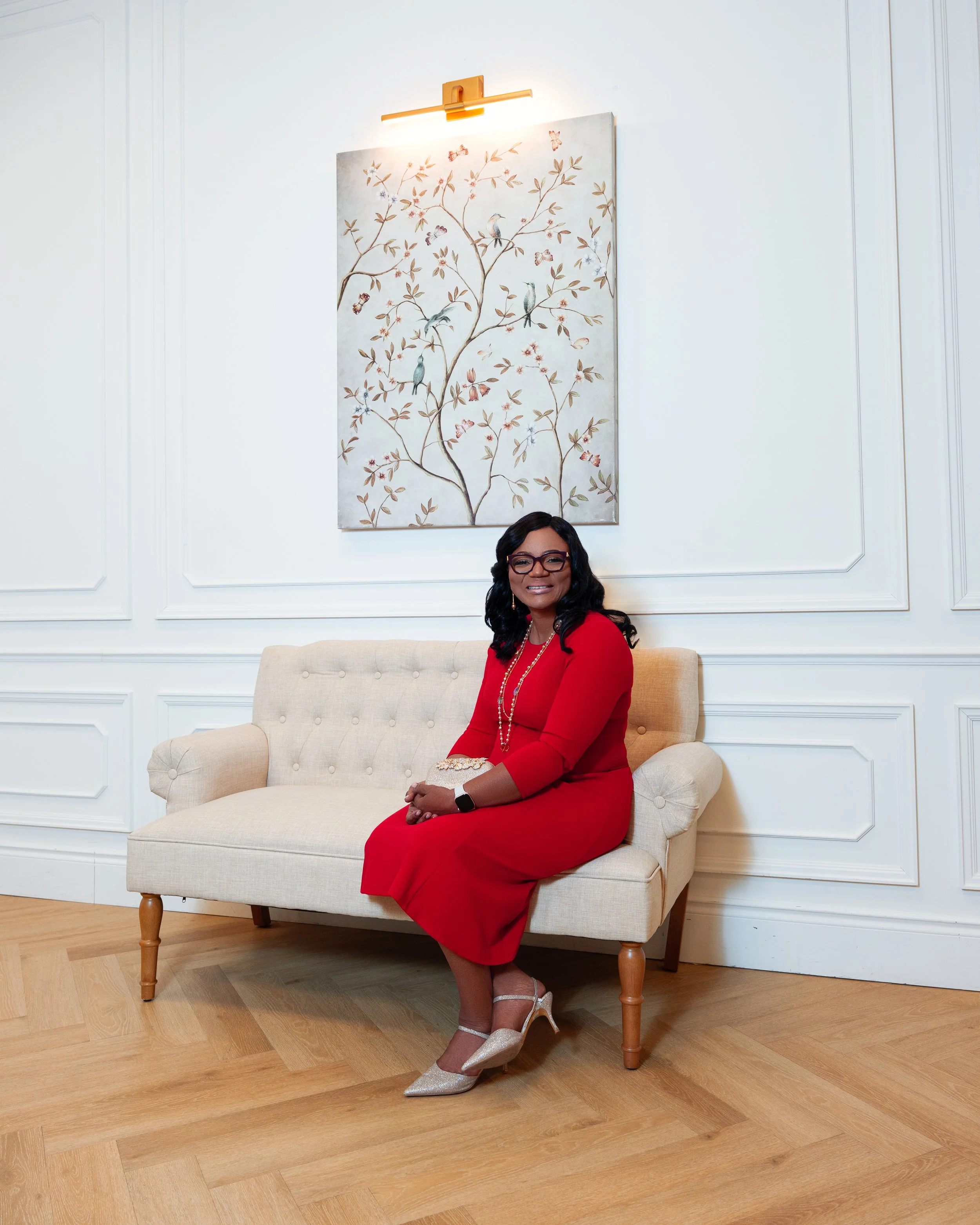 A woman wearing a red dress, beige high heels, earrings, glasses, and a watch sitting on a beige sofa in a room with white walls and a wooden floor, with a painting of a tree with birds, butterflies, and flowers hanging on the wall behind her, illumi