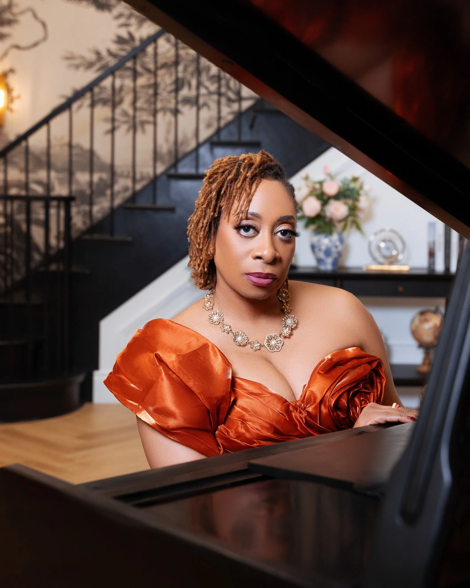A woman with styled hair wearing an orange dress and a jewelry necklace is playing the piano in a room decorated with a flower vase and picture frames on a white wall.