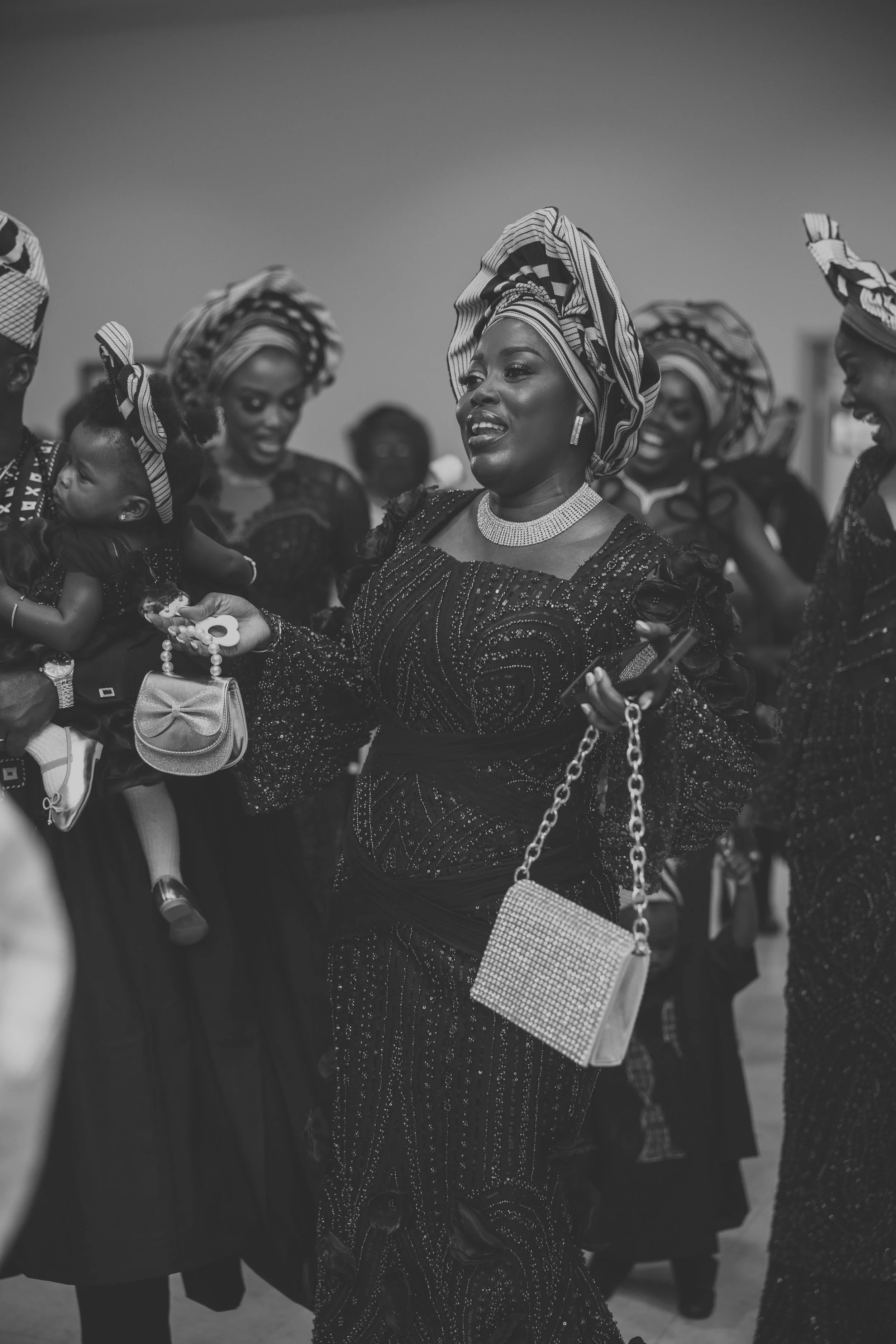 Women dressed in traditional African attire, including headwraps and jewelry, gathered together outdoors in a black and white photograph.