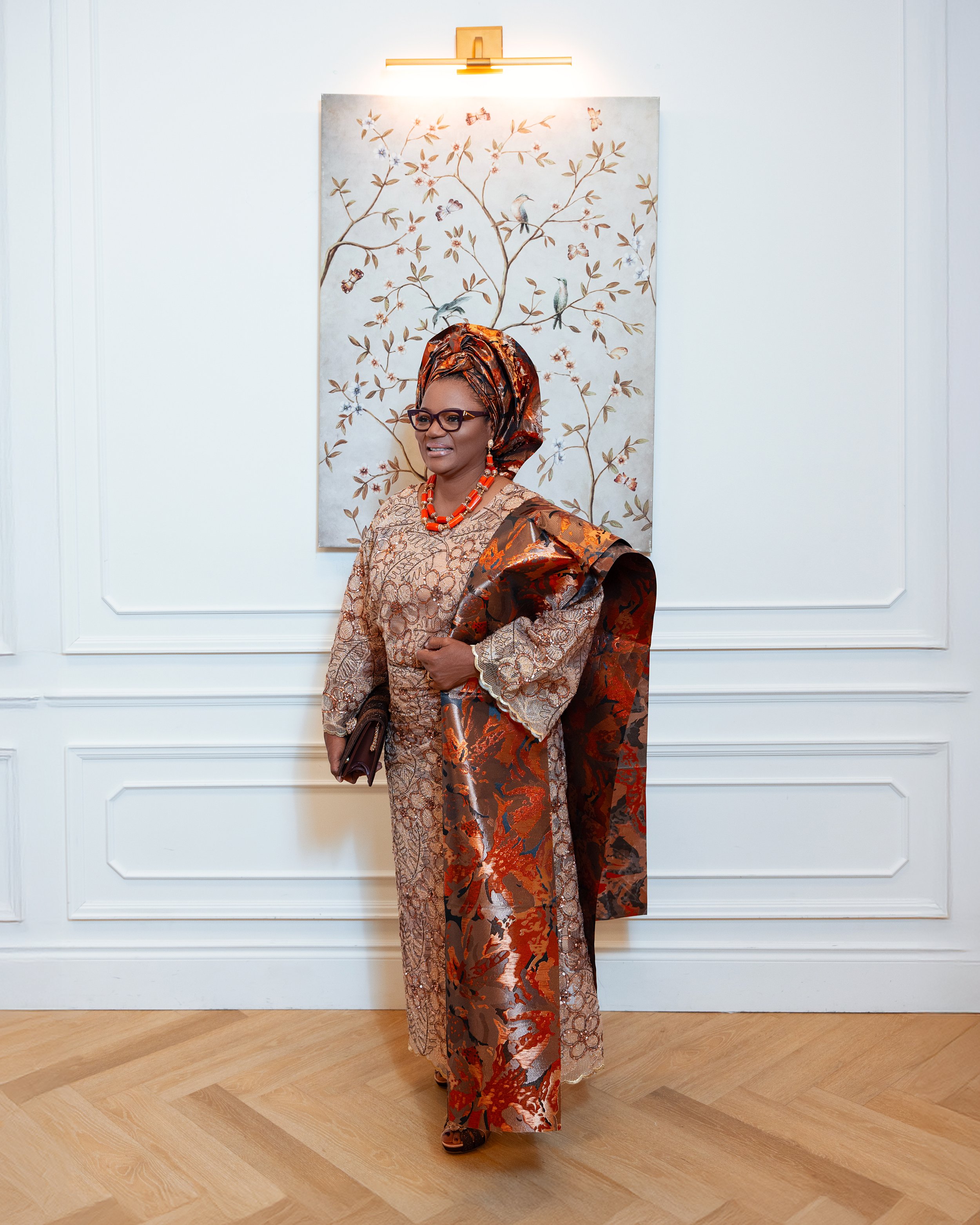 Woman dressed in traditional African attire standing in front of a decorative wall art of birds and butterflies on a white wall.