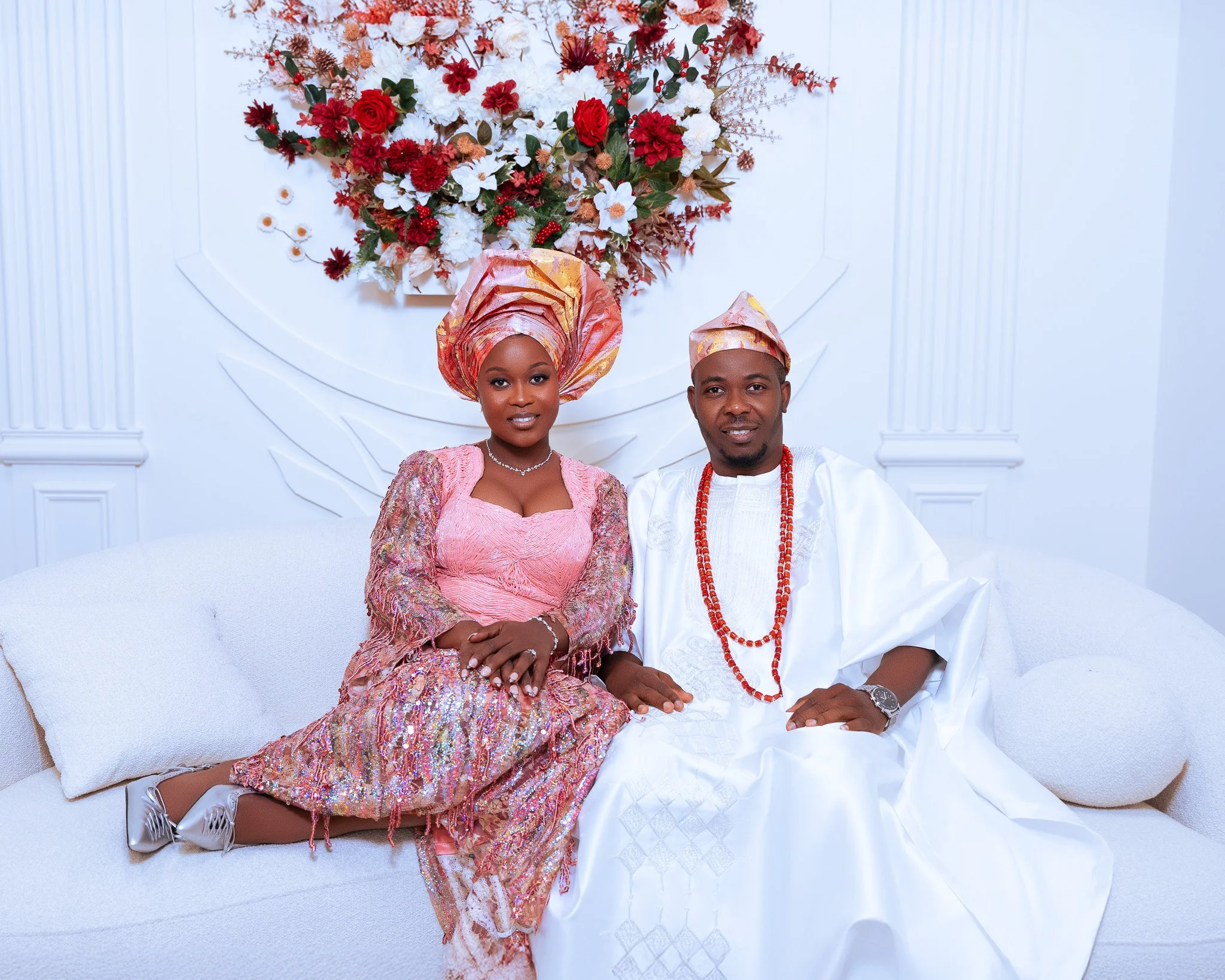 A woman and a man dressed in traditional African attire sitting on a white sofa with a large floral arrangement above them.