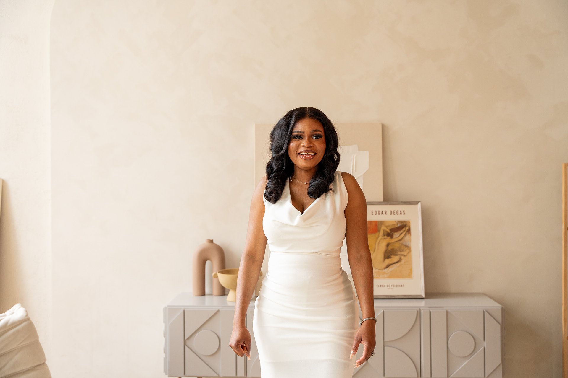 A woman with dark hair styled in loose curls is wearing a sleeveless white dress and smiling while standing in a minimalistic interior with neutral tones, behind her is a beige decorative vase, an abstract art poster, and a sideboard.