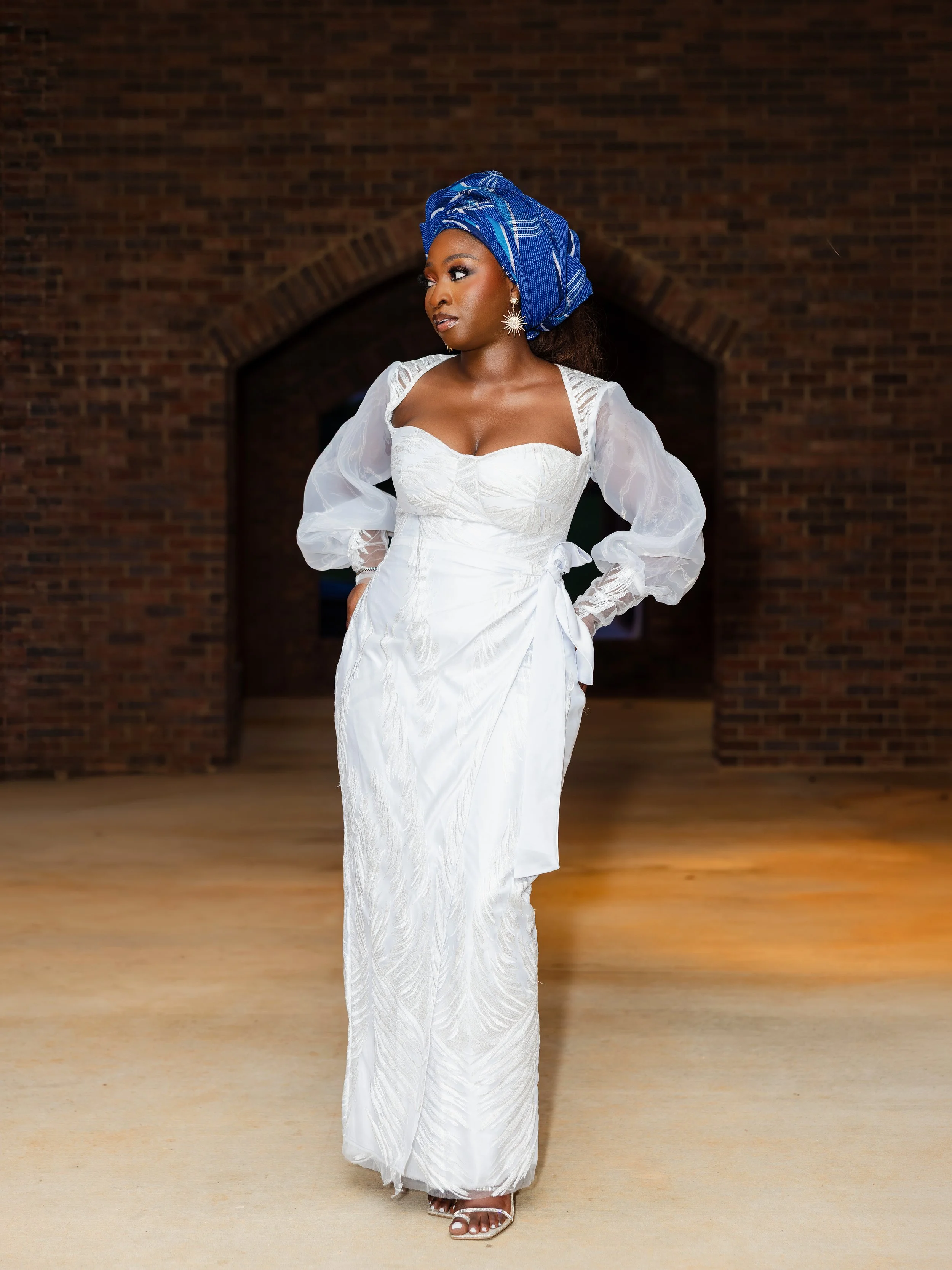 A woman in an elegant white dress with sheer puff sleeves and a fitted bodice, wearing a blue headwrap and star-shaped earrings, standing in front of a brick archway on a wooden floor.