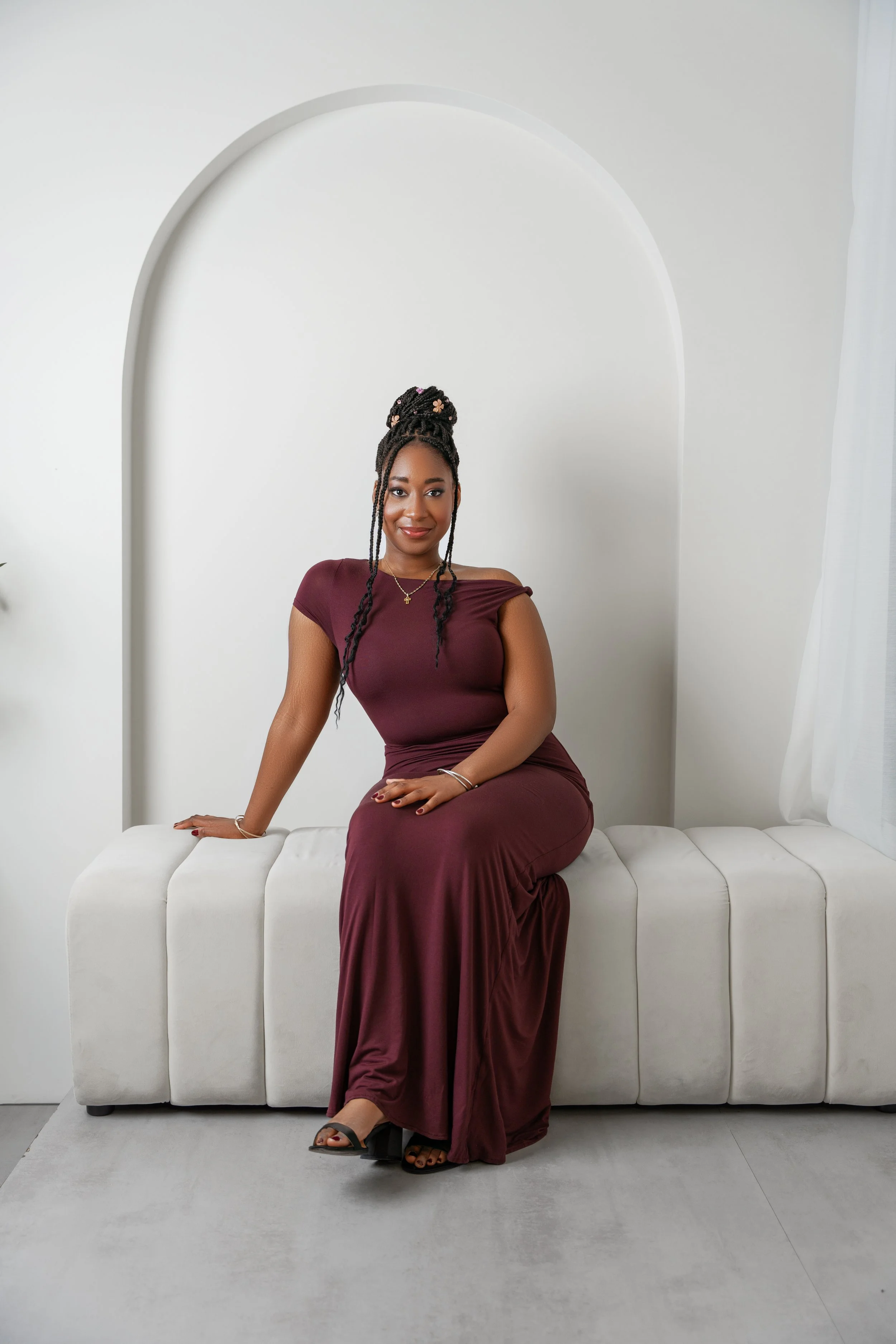 A woman with braided hair and a floral hair accessory, wearing a burgundy off-shoulder dress, sitting on a white tufted bench against a white wall with an arched design.