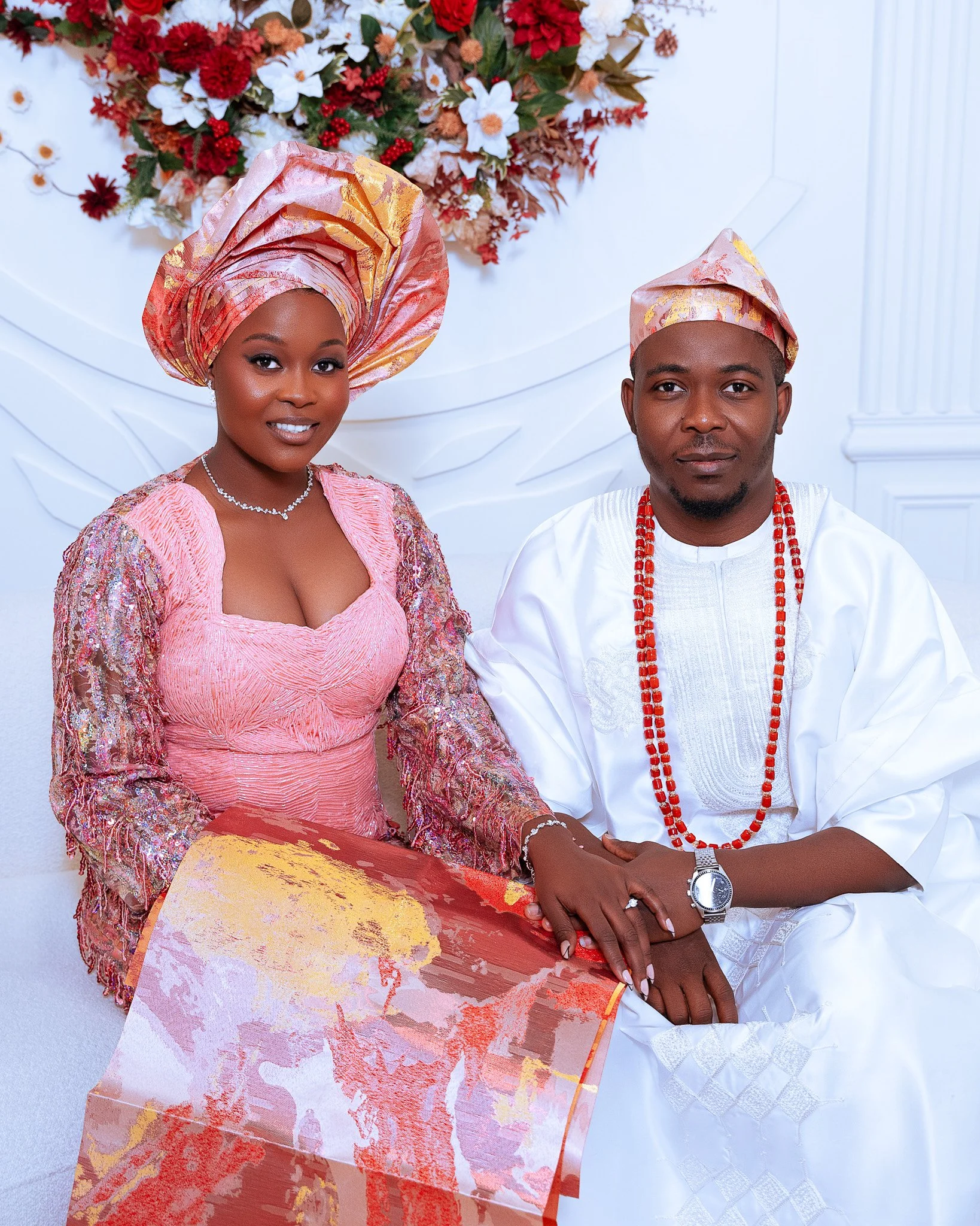 A couple dressed in traditional Nigerian attire, holding hands, with a floral backdrop in the background.
