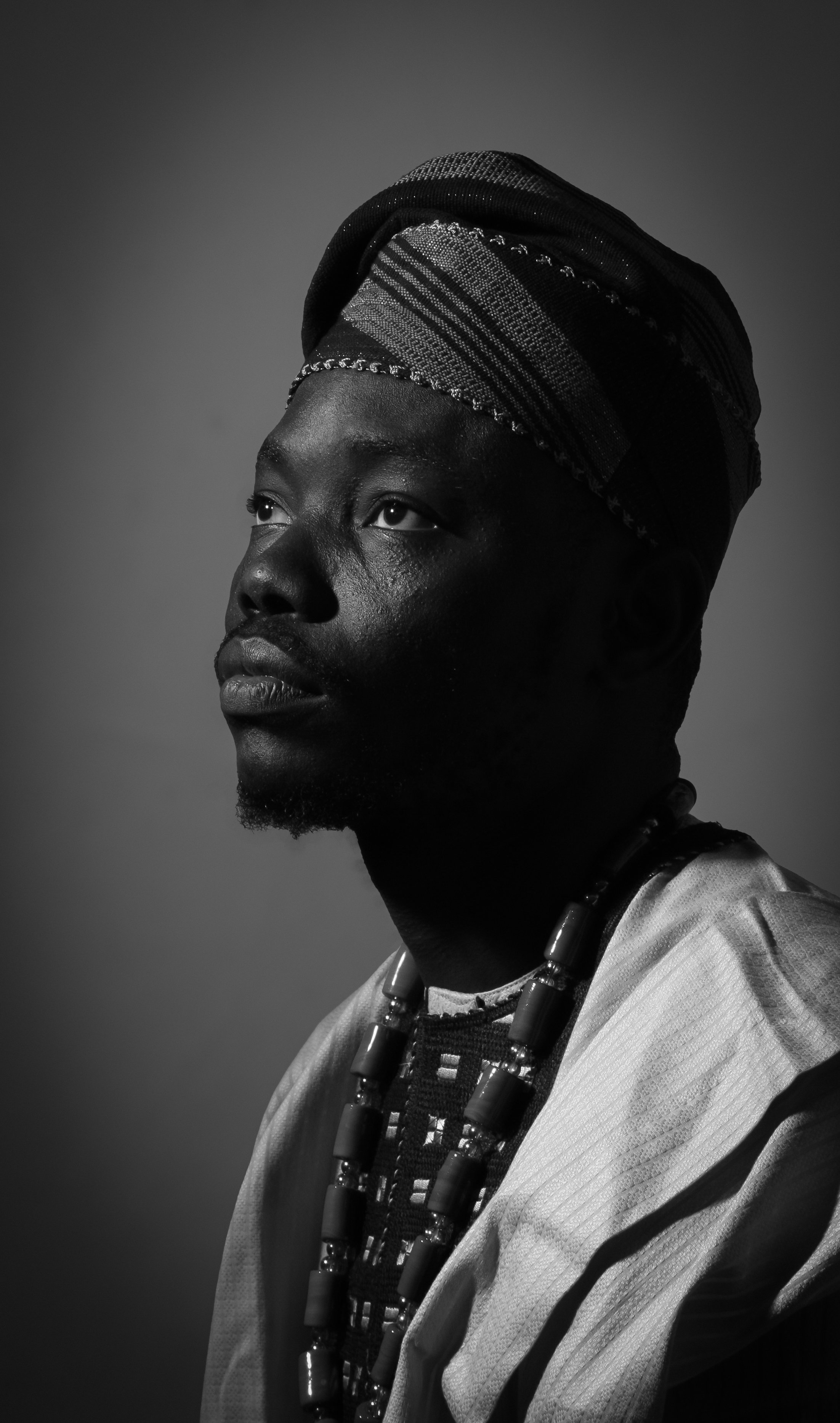 Black and white portrait of an African woman wearing traditional attire, including a headwrap and beaded jewelry, looking upwards.