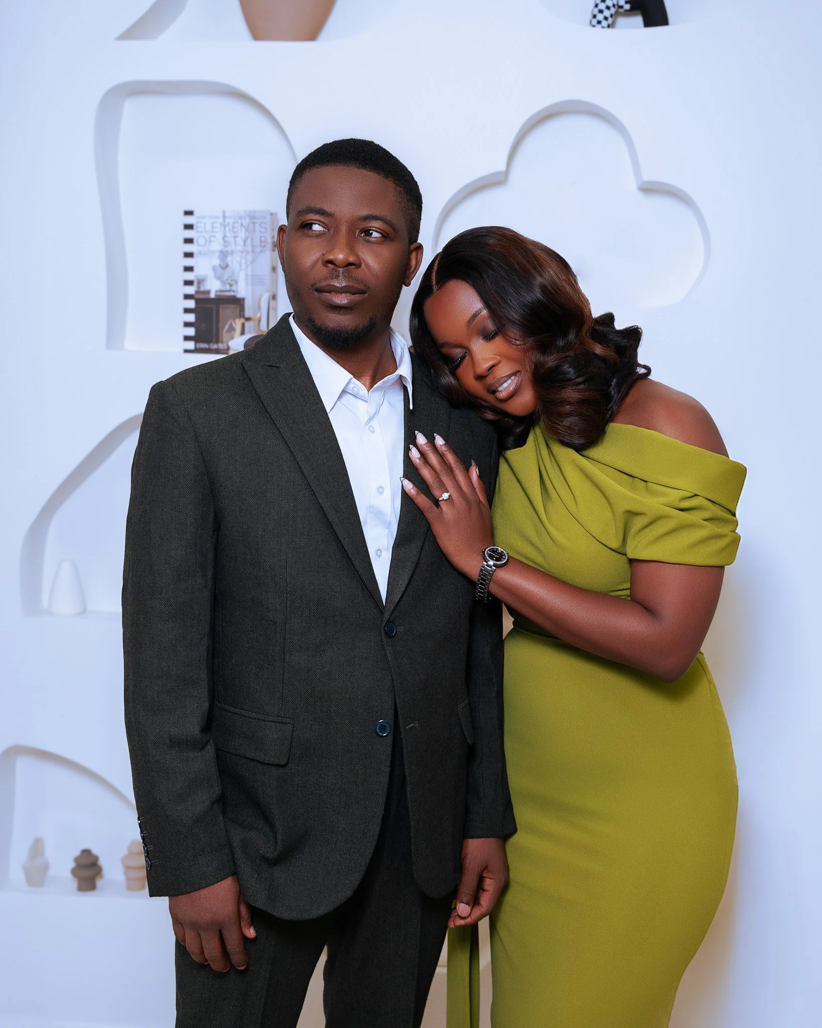 A man in a dark suit and a woman in a bright green dress standing close together, with the woman resting her head on the man's shoulder, in front of a white decorative backdrop.