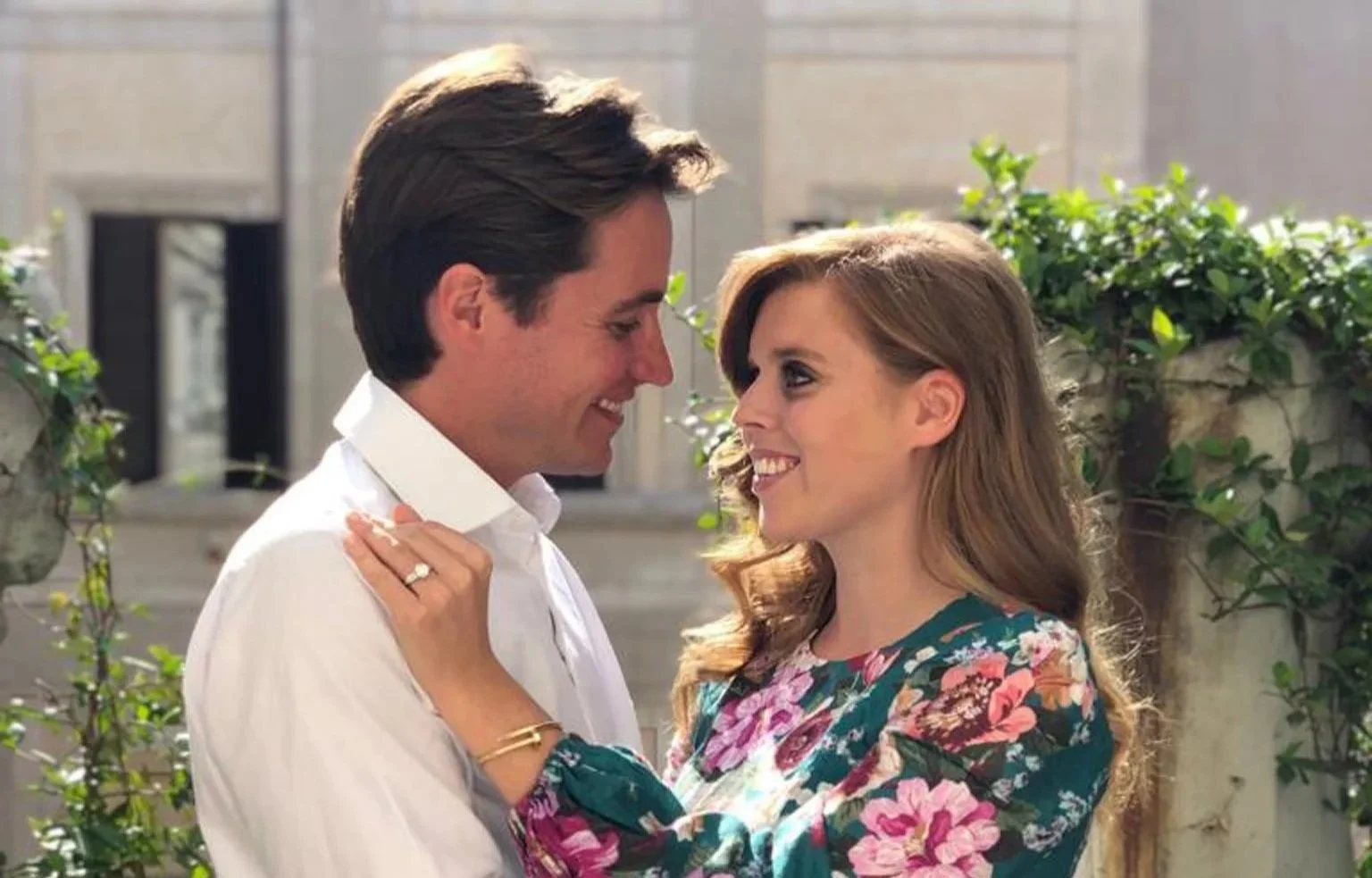 A man and a woman are standing close, smiling, and looking into each other's eyes outdoors. The man has dark hair and is wearing a white shirt, while the woman has light brown hair and is dressed in a floral top. They appear happy and affectionate.