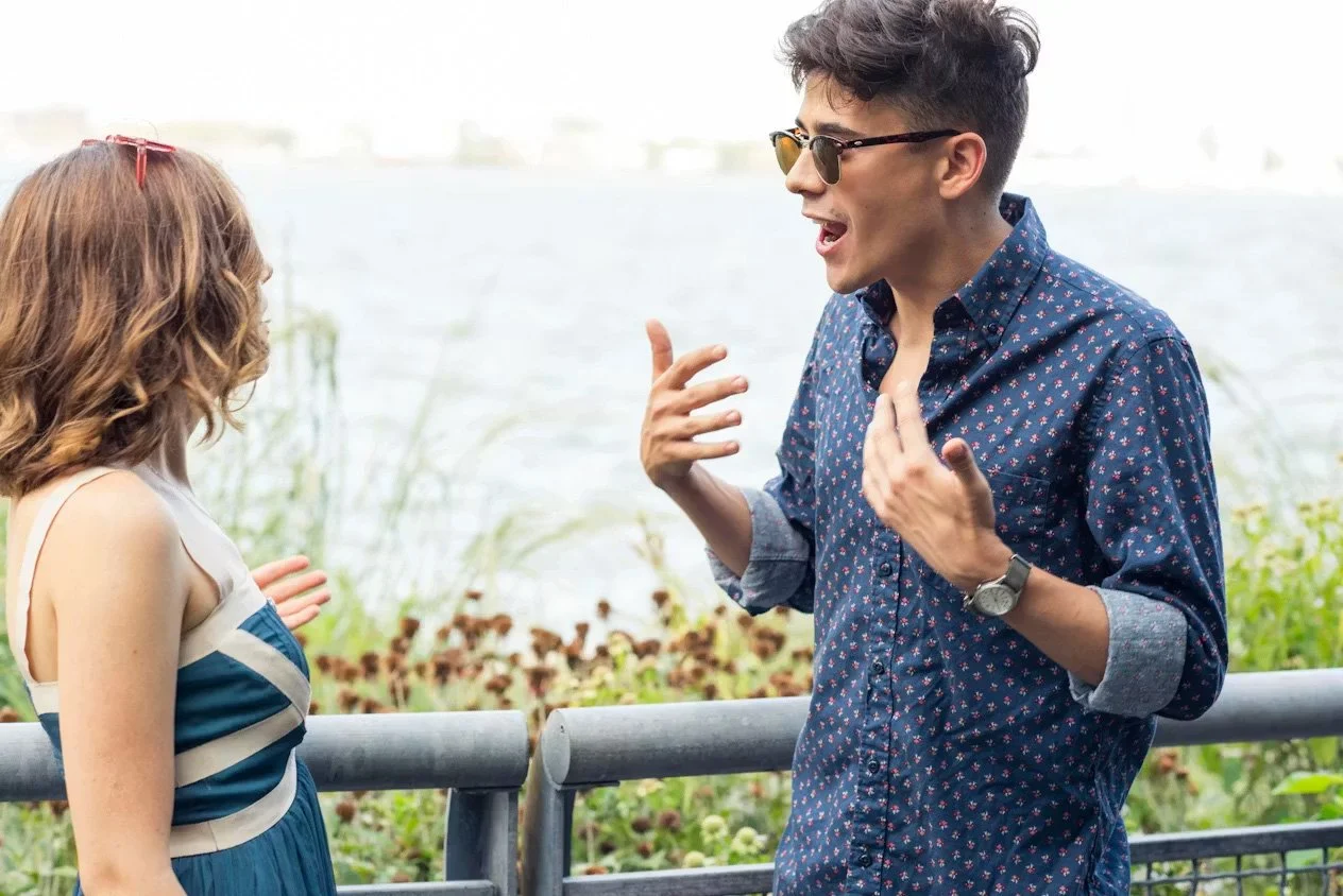 A young man and woman having a conversation outdoors by a body of water. The man is gesturing with his hands, wearing glasses and a patterned shirt. The woman has shoulder-length hair and is wearing sunglasses on her head and a blue and white striped dress.