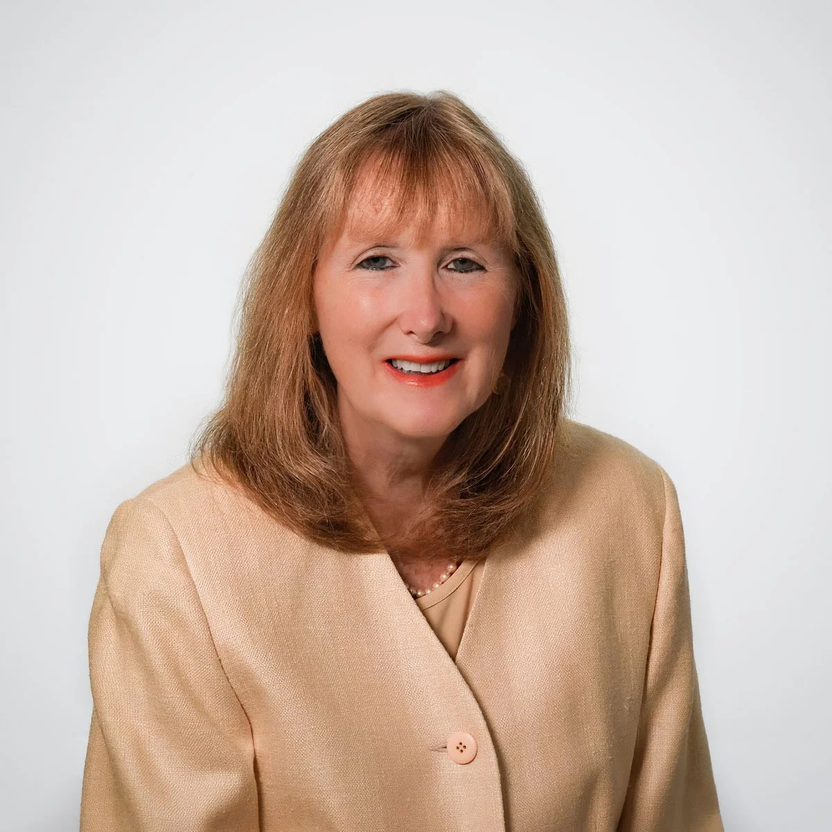 Photograph of a middle-aged woman with shoulder-length light brown hair, wearing a beige blazer and a pearl necklace, smiling against a plain white background.