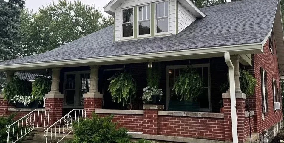 Front view of a brick house with a porch, hanging plants, and a gabled roof.