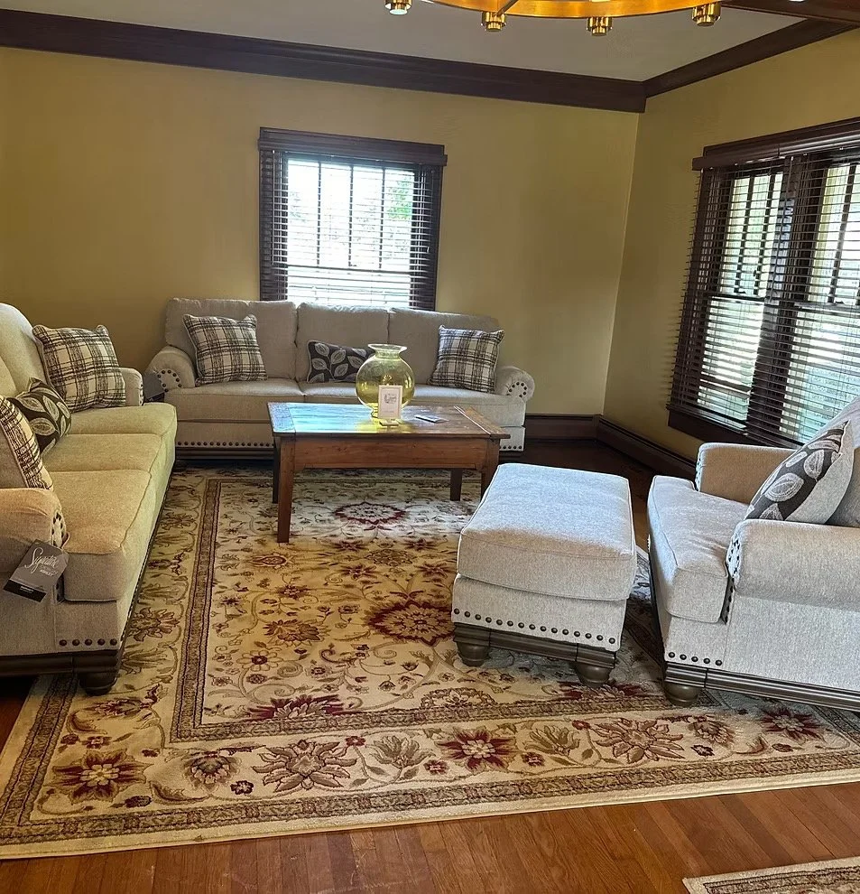 Living room with three sofas, a wooden coffee table with a glass vase, a patterned area rug, and windows with wooden blinds.