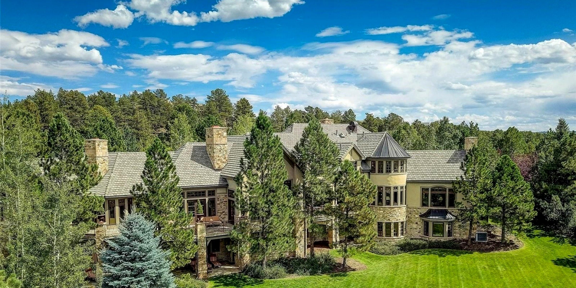 A large house with a stone and stucco exterior, multiple chimneys, and a tiled roof, surrounded by tall trees and a well-maintained lawn under a partly cloudy sky.