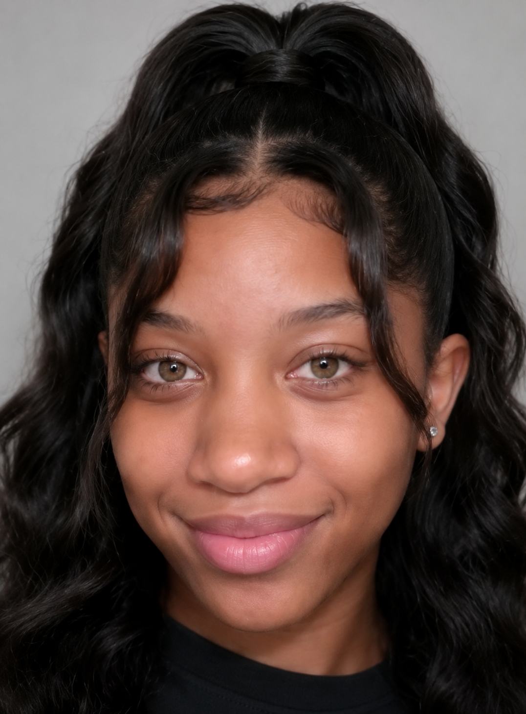 Close-up of a young woman with wavy black hair and light brown eyes, smiling softly, wearing a black top and small earrings, against a neutral background.