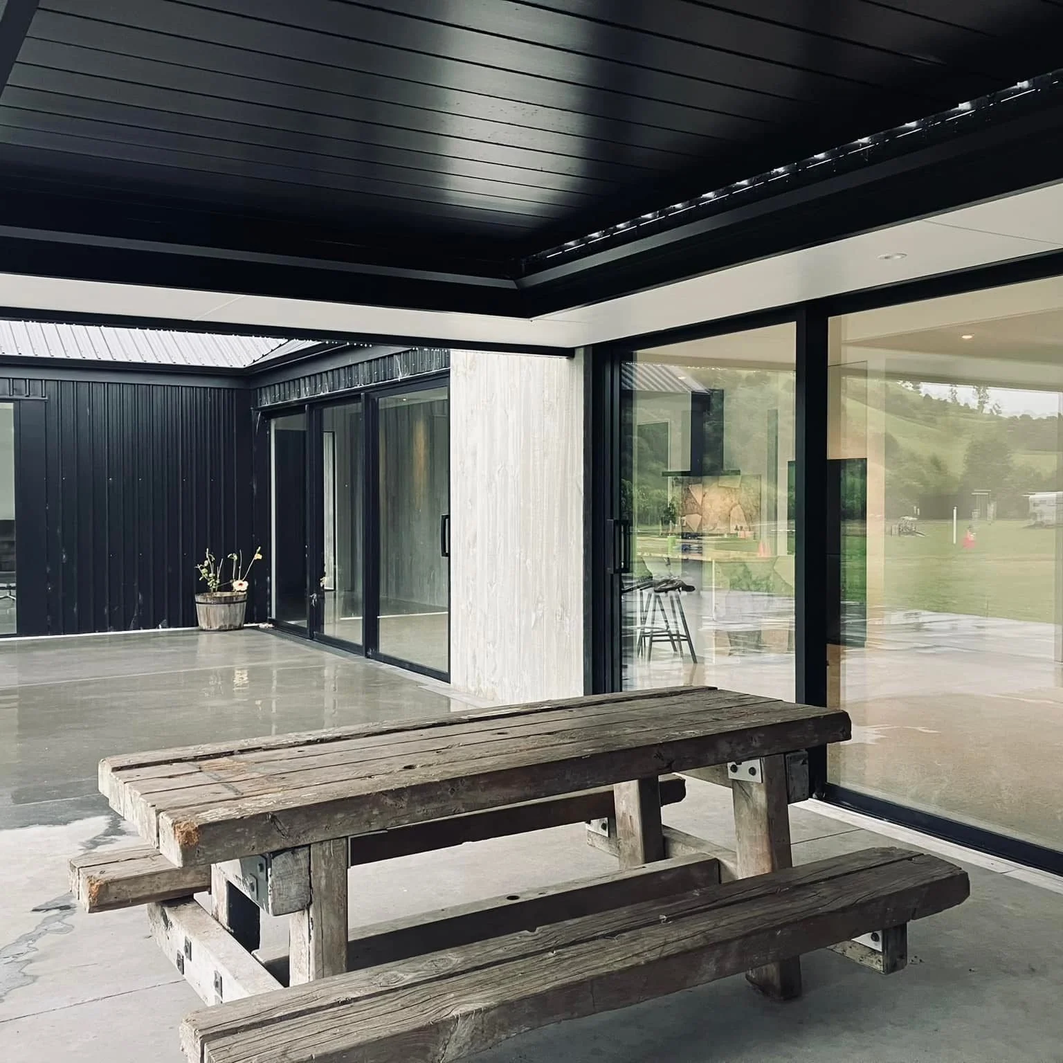 View of an outdoor patio area with a wooden picnic table, concrete flooring, and a modern house with large glass sliding doors, black framing, and a black ceiling.