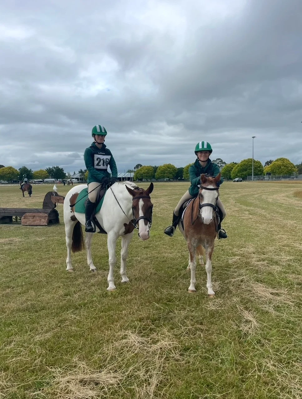 Two children on horseback riding on a grassy field under cloudy skies, wearing green helmets and riding attire.