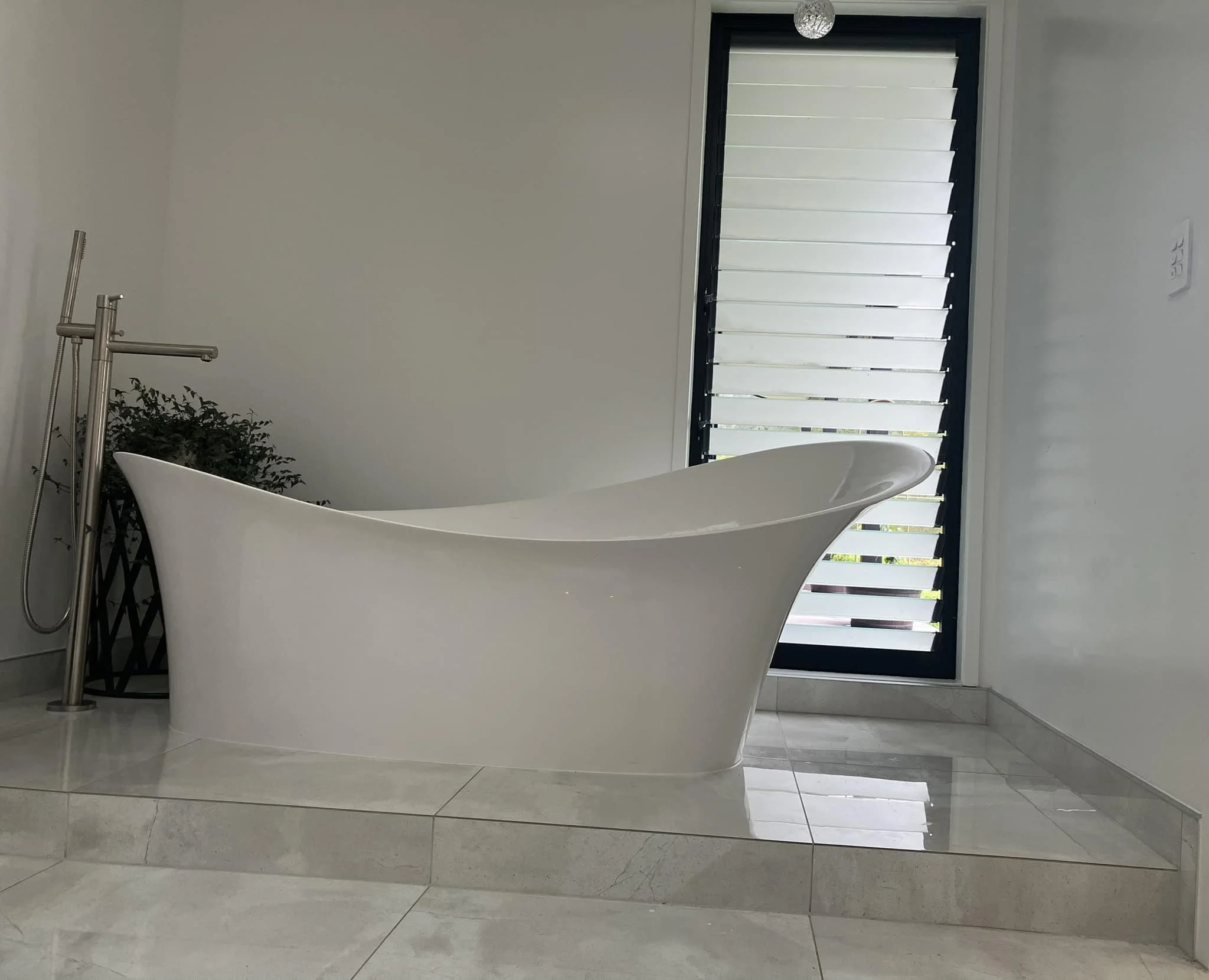 Modern bathroom with a white freestanding bathtub, large gray tiled floor, and a window with black horizontal blinds.