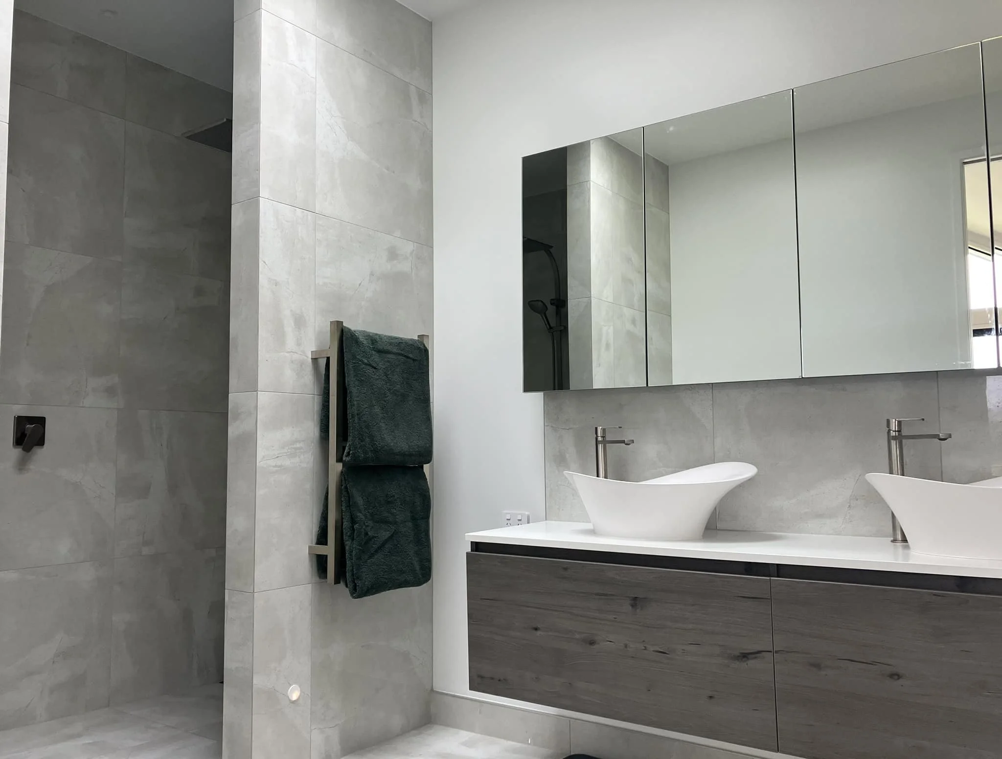Modern bathroom with gray tile walls, a large mirror, a white vessel sink, and a wooden cabinet below the sink. There is a towel hanging on a ladder-style towel rack, and a window is visible with partial sunlight.