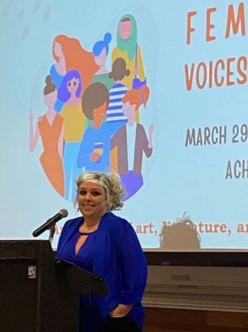 A woman in a blue blouse standing behind a podium with a microphone at a Women's Voices event on March 29.