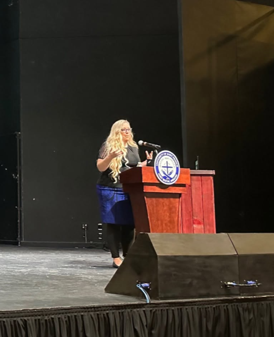 Woman with long blonde hair and glasses speaking at a podium on a stage with a black wall background.
