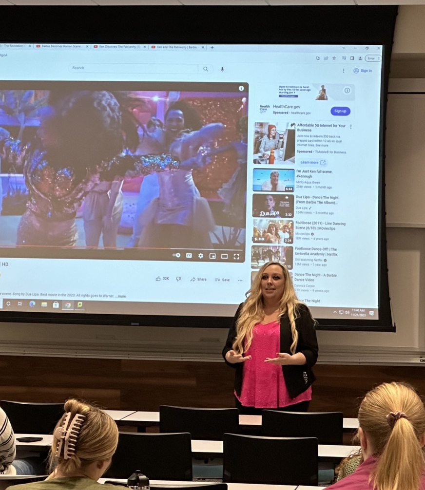 A woman with long blonde hair giving a presentation in a classroom with students. A large screen behind her displays a YouTube video paused on a scene from the movie, Barbie.