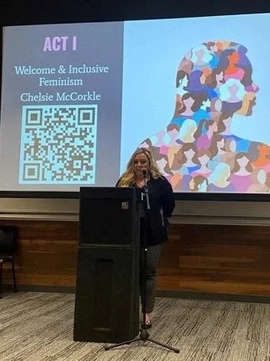 A woman standing behind a podium giving a presentation in a conference room. A large screen behind her displays a colorful illustration of diverse women, along with the text 'ACT I Welcome & Inclusive Feminism Chelsie McCorkle' and a QR code.