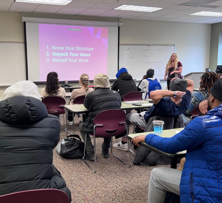 Classroom with students seated at desks, listening to a female teacher standing near a whiteboard. A PowerPoint slide titled '1. Know Your Structure, 2. Unpack Your Ideas, 3. Reread Your Work' is projected on the screen.