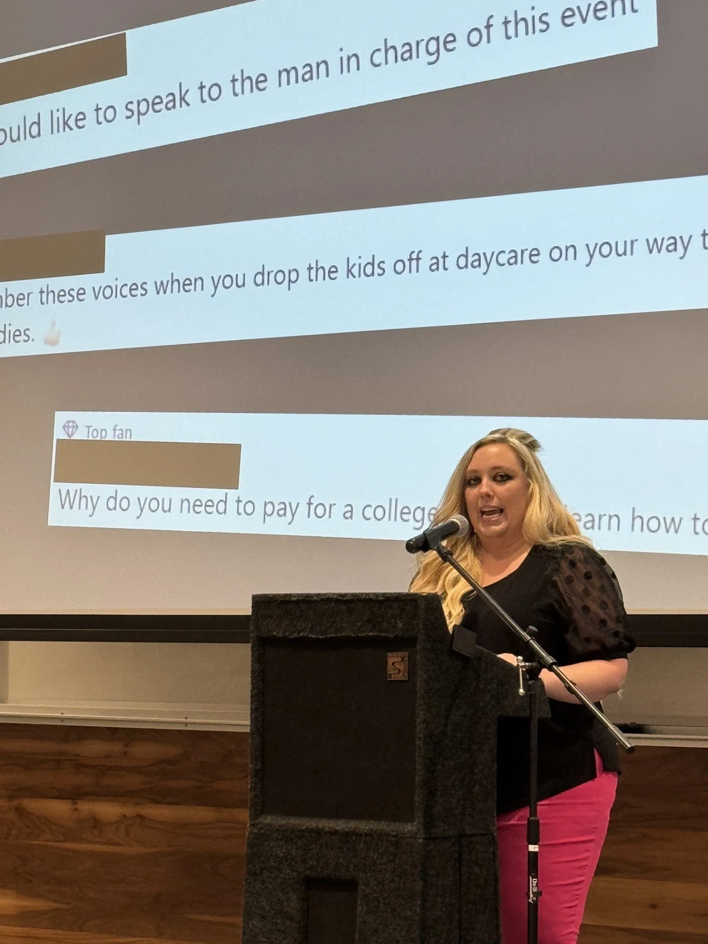 A woman with blonde hair and a black top speaking at a podium, with a large projected presentation behind her. The presentation has text on a white background with brown accents.