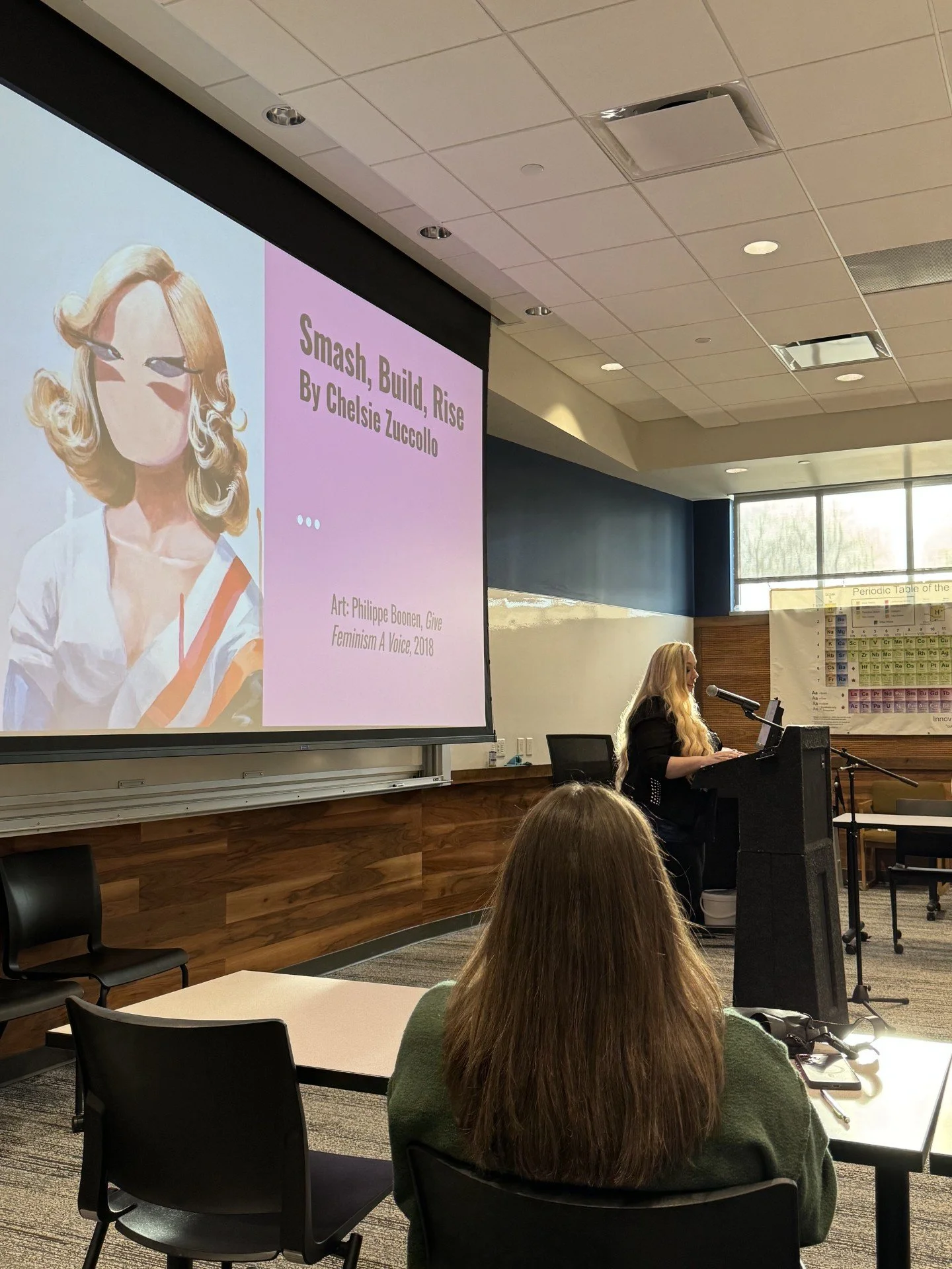 A woman in a classroom or lecture hall with a large presentation slide displayed on a screen behind her. The slide features an artistic portrait of a woman with blonde, curly hair and the title 'Smash, Build, Rise' by Chelsie Zuccolo, along with artistic credits.