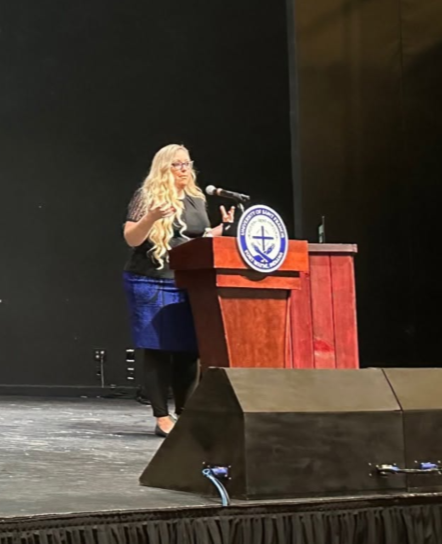 A woman with long blonde hair, wearing glasses, a black top, and a blue skirt, standing at a wooden podium with a microphone, speaking to an audience in an auditorium.