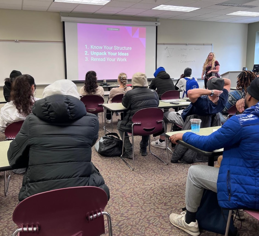 A classroom with students seated at desks listening to a female instructor standing near a whiteboard. A large screen displays a presentation with three points: 'Know Your Structure,' 'Unpack Your Ideas,' and 'Reread Your Work.'