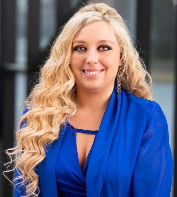 A woman with long, blonde, curly hair smiling in front of a glass background, wearing a blue blouse or dress.
