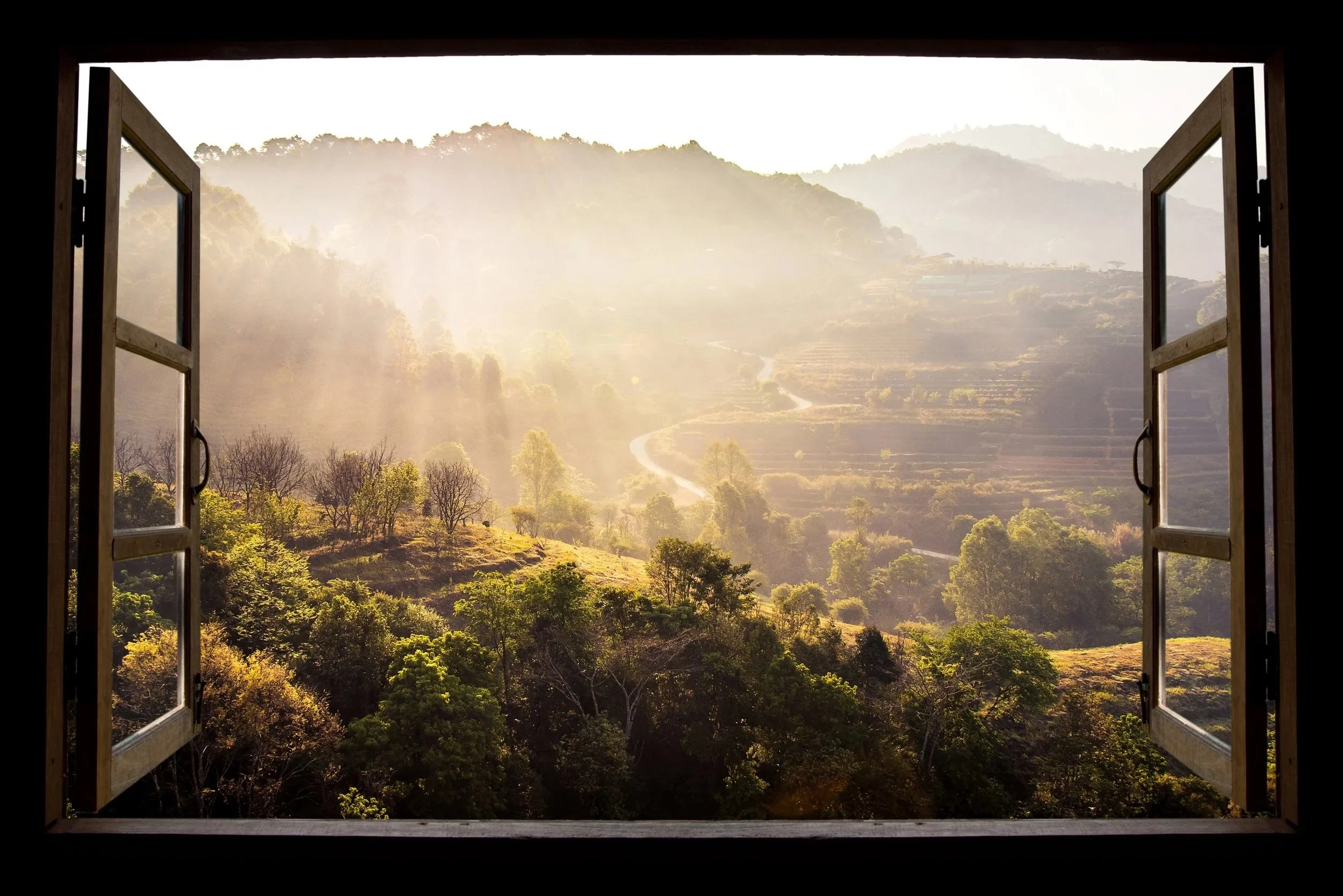 Open window overlooking misty, tree-covered mountains with winding road in the valley.