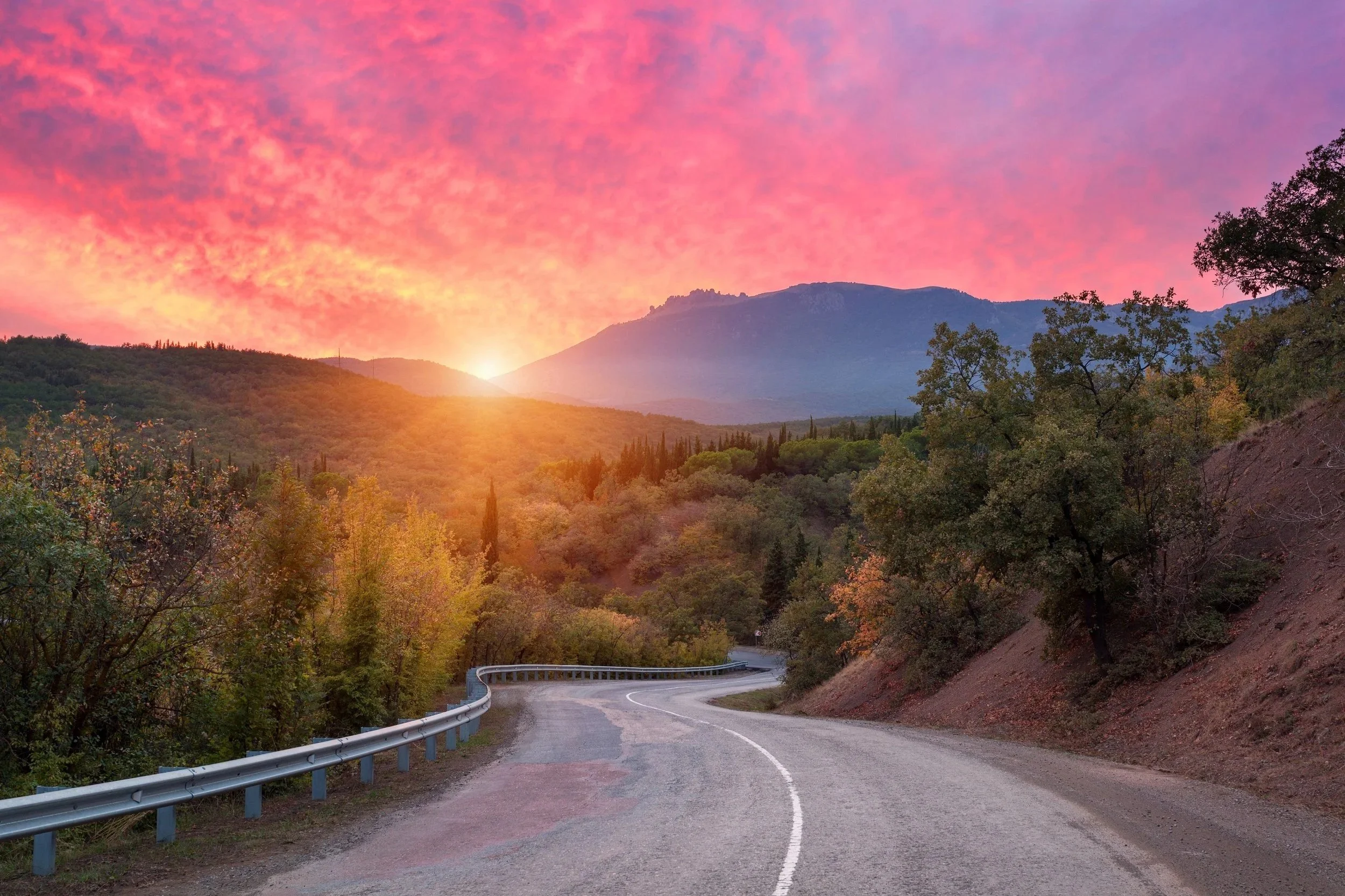 A winding mountain road at sunset, with a guardrail on the left, trees on both sides, and mountains in the background under a pink and orange sky.