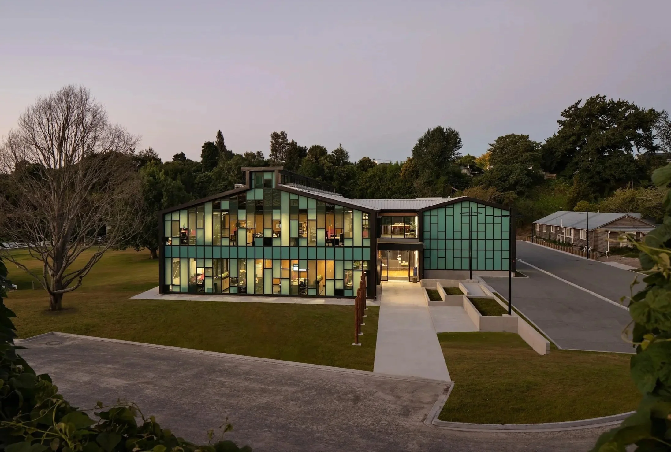 The Kollective, Tauranga. Modern glass building with illuminated interior, surrounded by trees and a paved driveway in the evening. Crowther & Company. Quantity surveyors and cost consultant specialists.