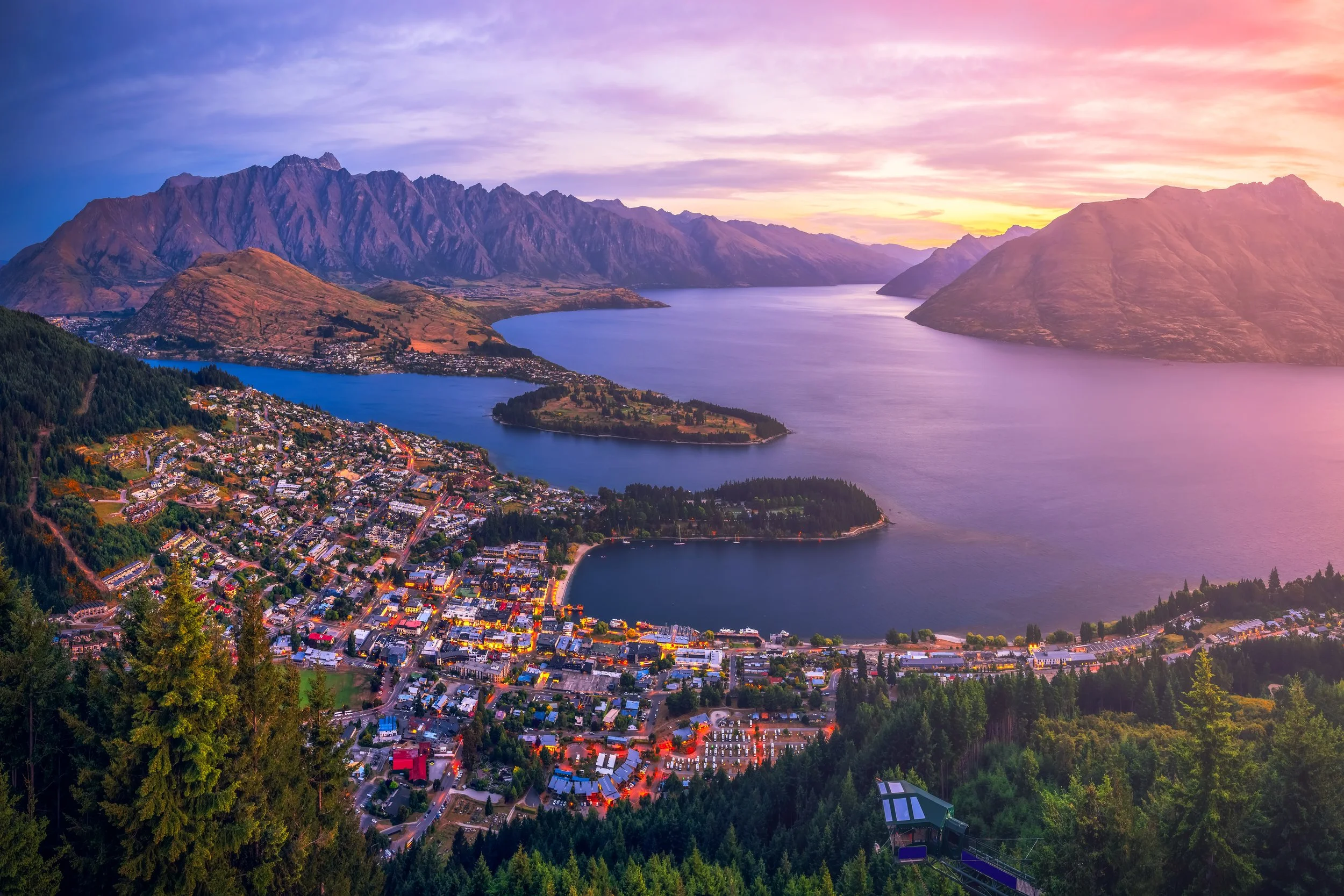 A scenic landscape of a town by a lake surrounded by mountains during sunset, with colorful sky and lush green trees in the foreground.