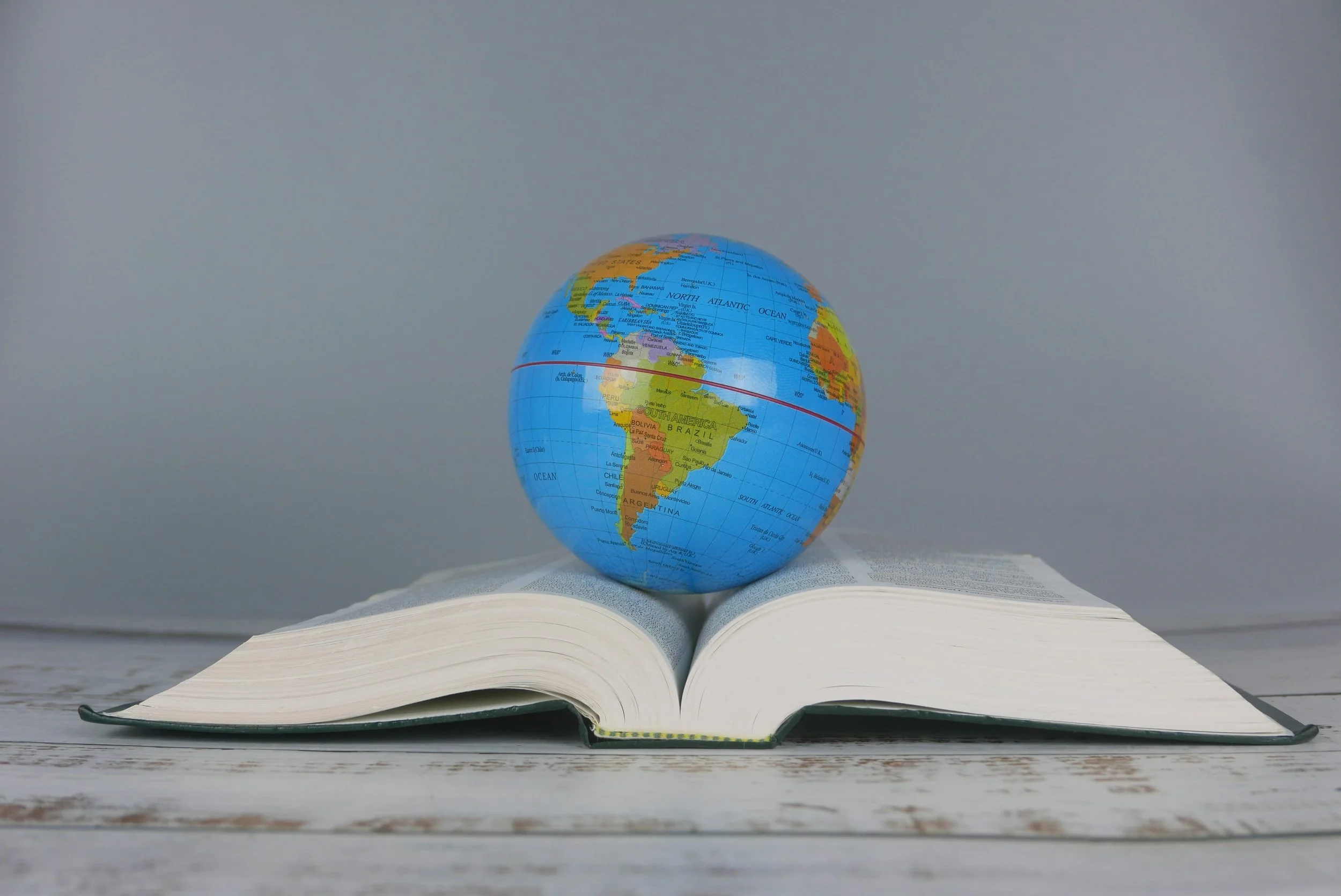 A globe sitting on top of an open book, placed on a white wooden surface with a gray background.