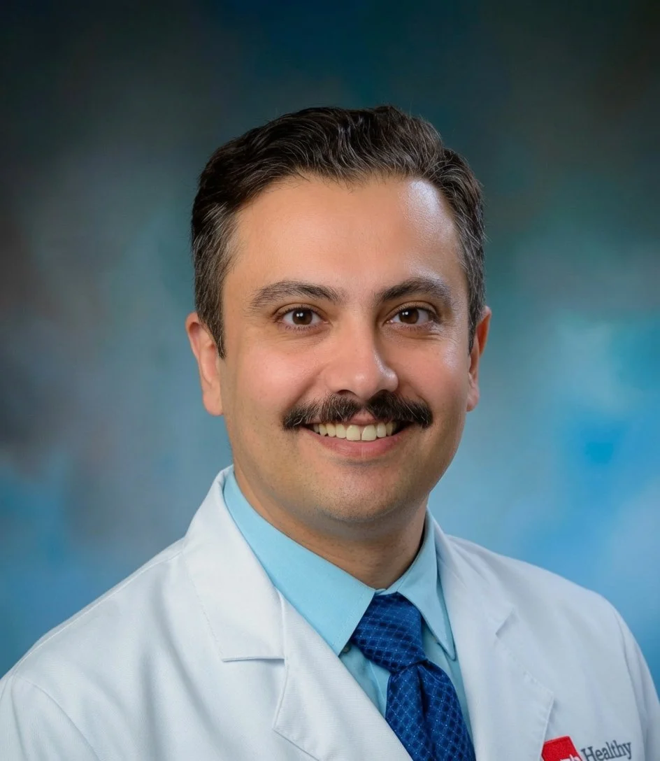 Portrait of a smiling male doctor with dark hair, a mustache, wearing a white lab coat, light blue shirt, and dark blue tie, against a blue background.