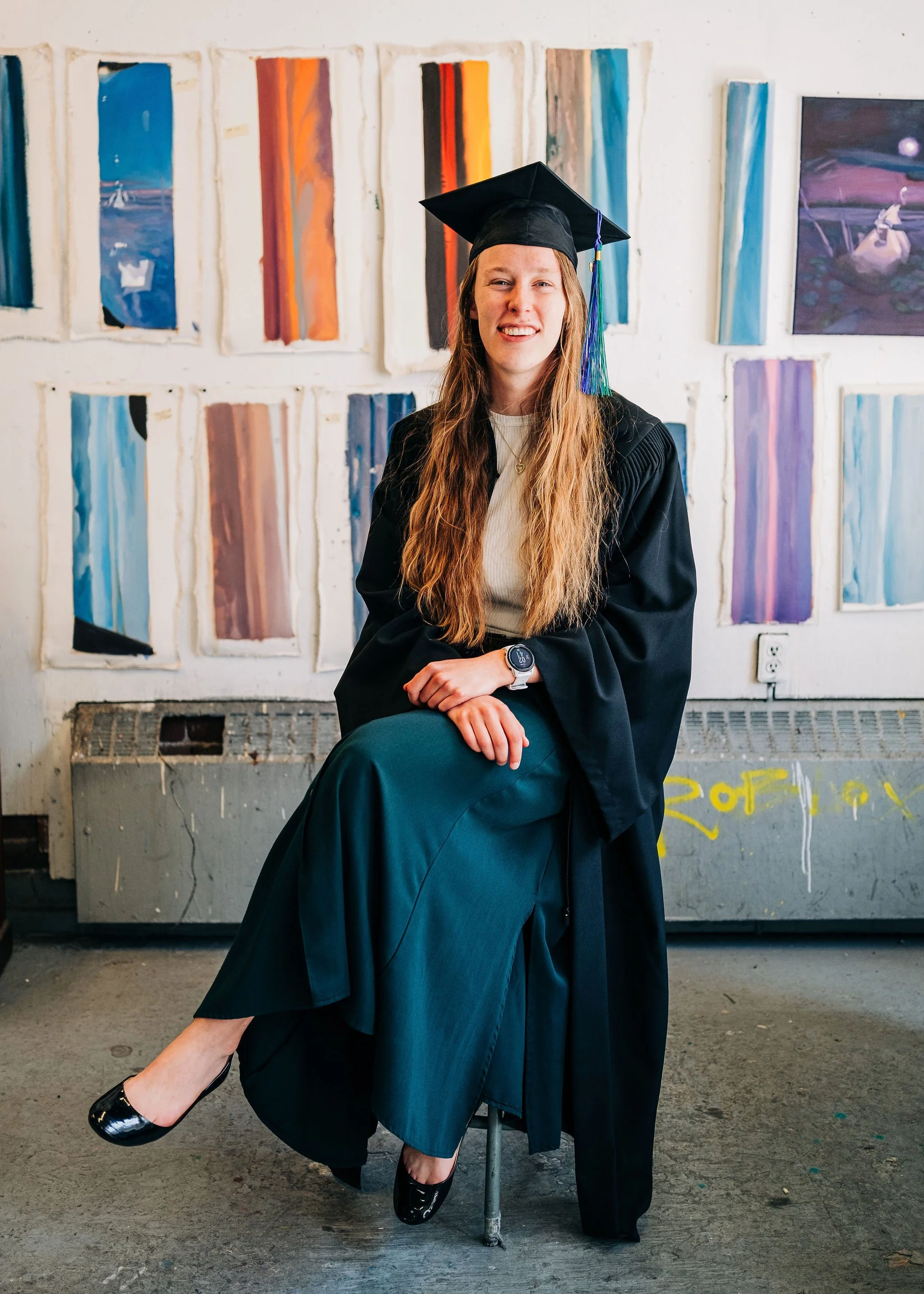 A young woman in a graduation cap and gown sitting on a stool with one leg crossed over the other. She is smiling and has long, wavy, light brown hair. The background features a white wall with colorful abstract paintings.