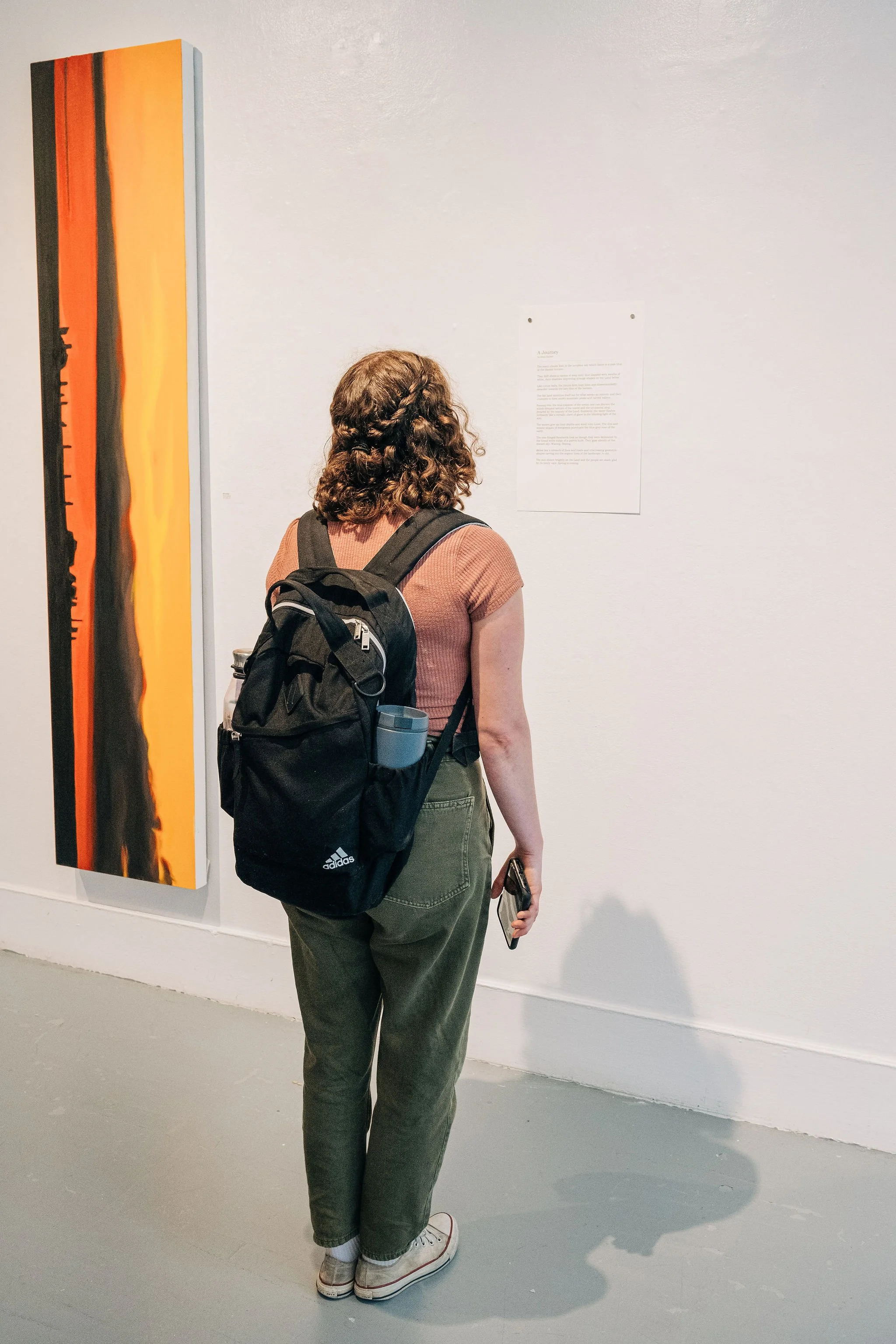 A woman with curly brown hair, wearing a brown shirt, green cargo pants, and white sneakers, stands with her back to the camera, carrying a black backpack and holding a smartphone, while observing an art piece on a gallery wall.
