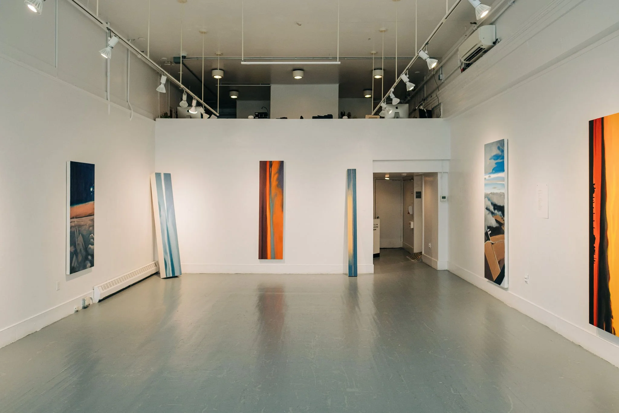 An art gallery with four vertical abstract paintings on white walls, featuring swirling blue, orange, and black colors. The gallery has a gray floor and a high ceiling with focused lighting.