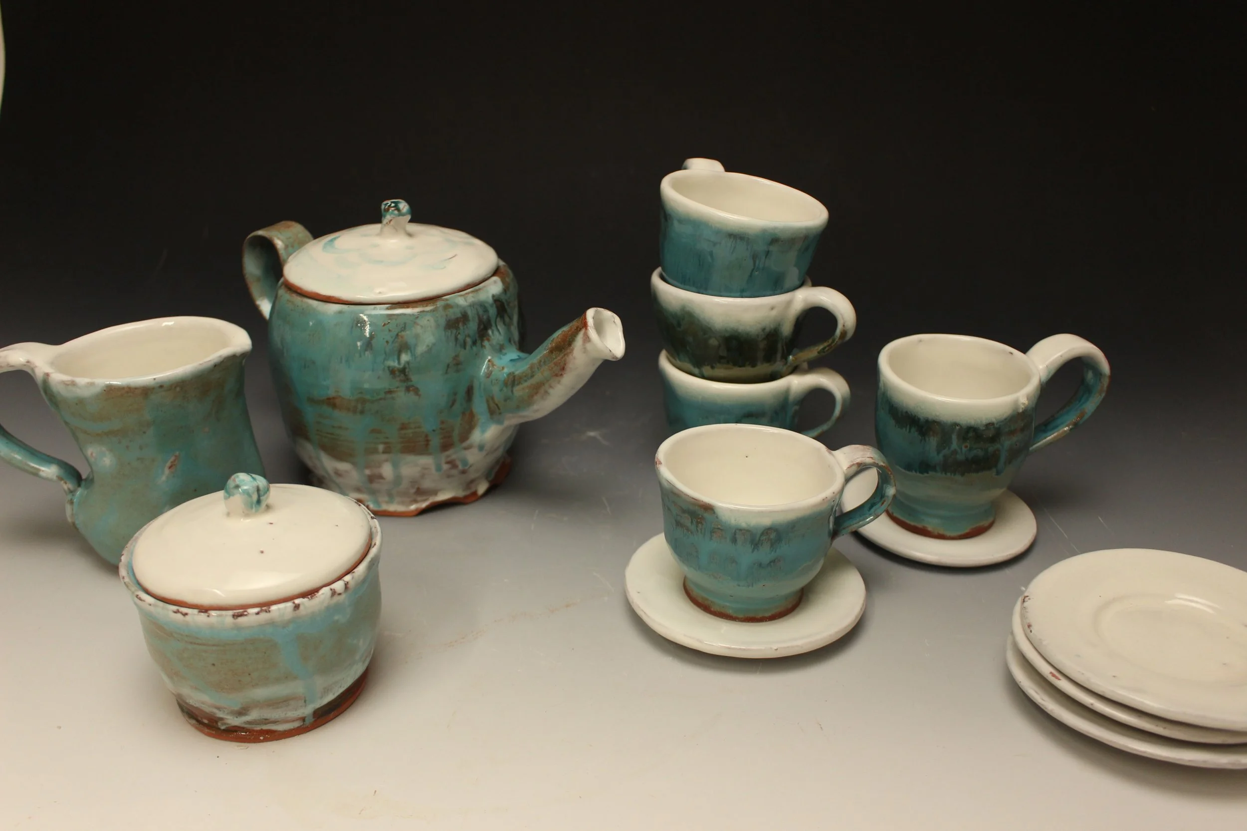 A collection of ceramic tea cups, a teapot with a lid, and matching saucers, all with a blue and white glaze design, arranged on a surface against a dark background.