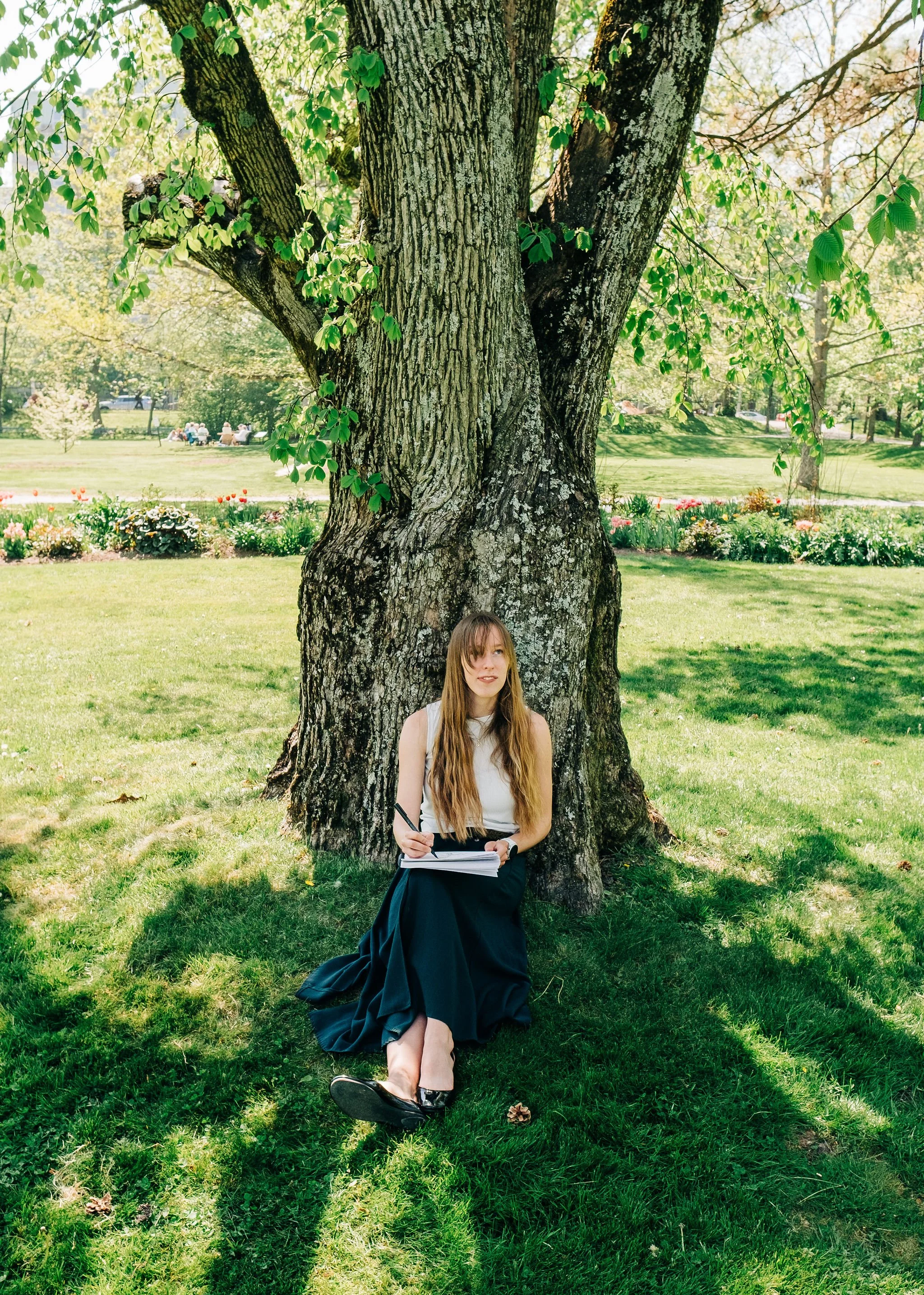 A young woman with long hair, wearing a sleeveless top and a long dark skirt, sitting under a large tree in a park on a sunny day with green grass and colorful flowers in the background.