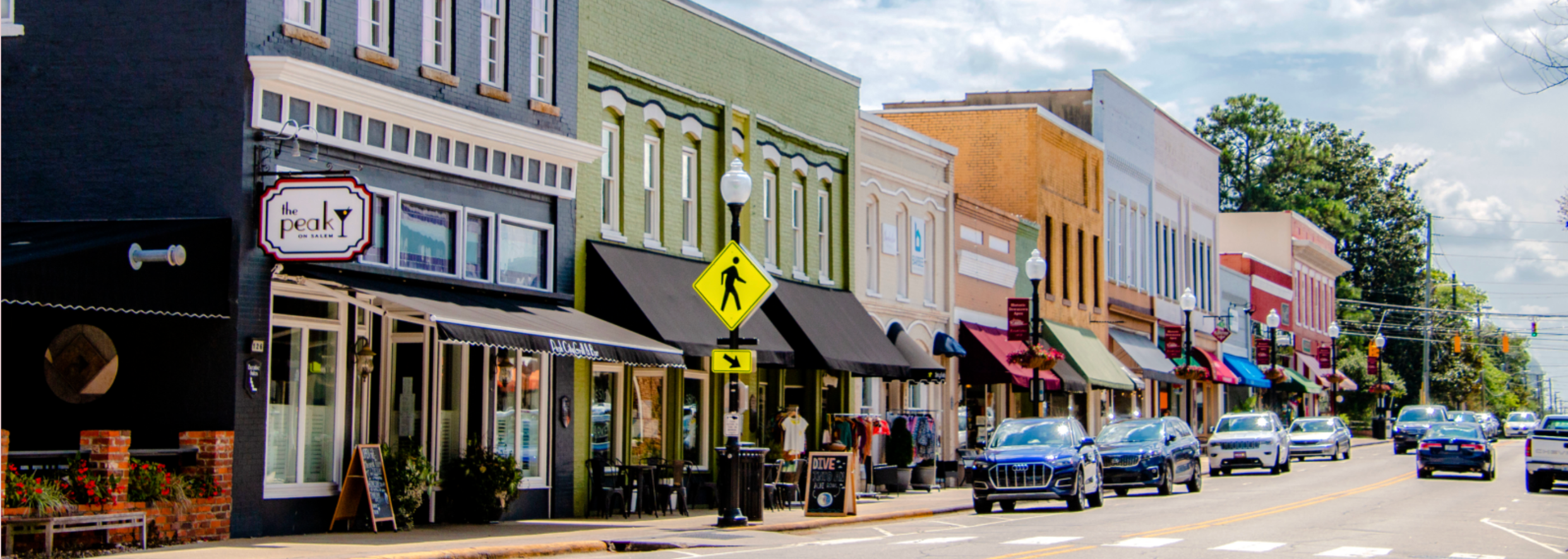 Colorful storefronts on a small town street with parked cars, pedestrian crossing sign, and trees in the background.