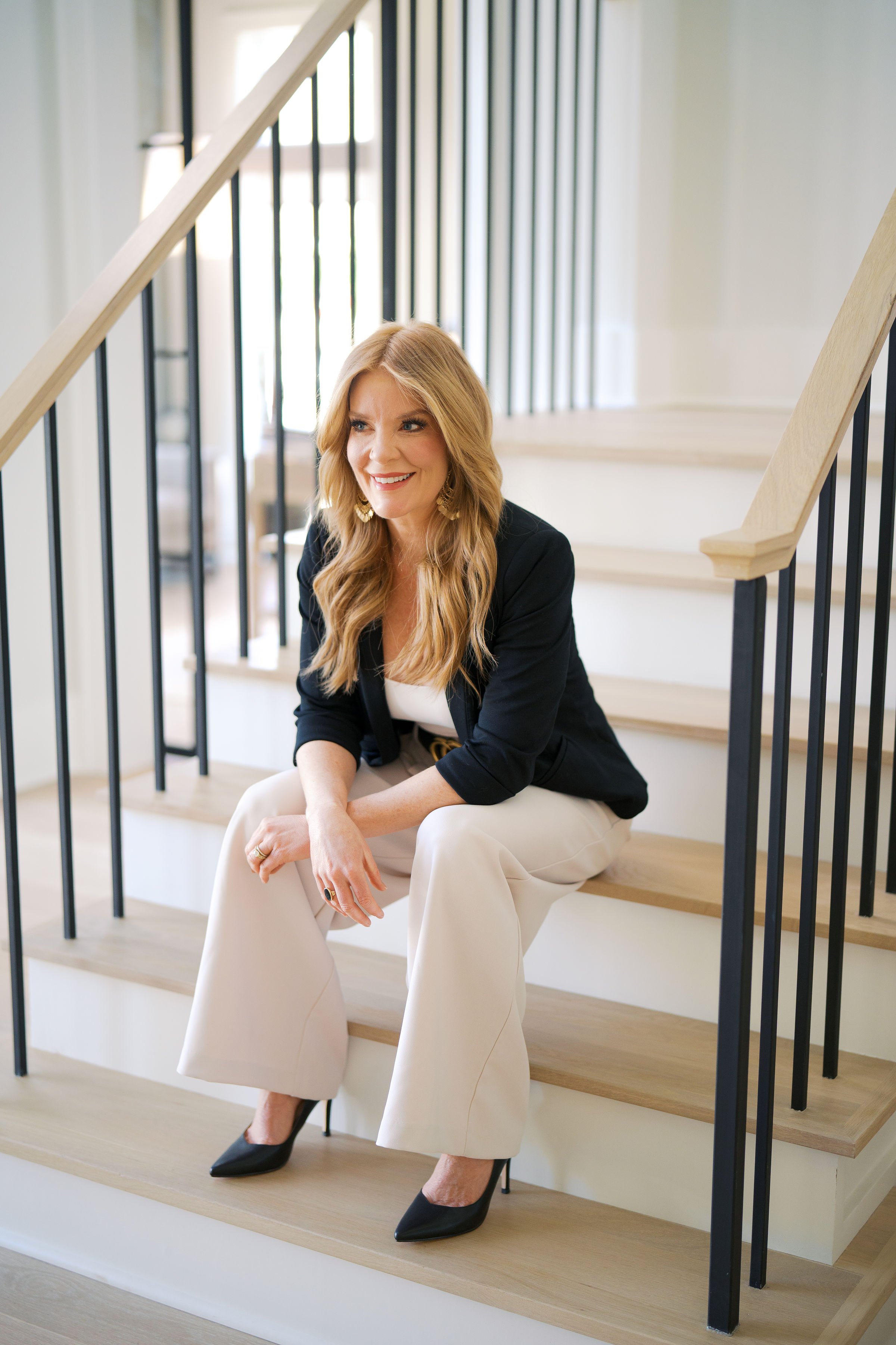A woman with wavy blonde hair sitting on stairs indoors. She is smiling, wearing a black blazer, white top, beige wide-leg pants, and black pointed shoes, with a ring on her finger and gold earrings.