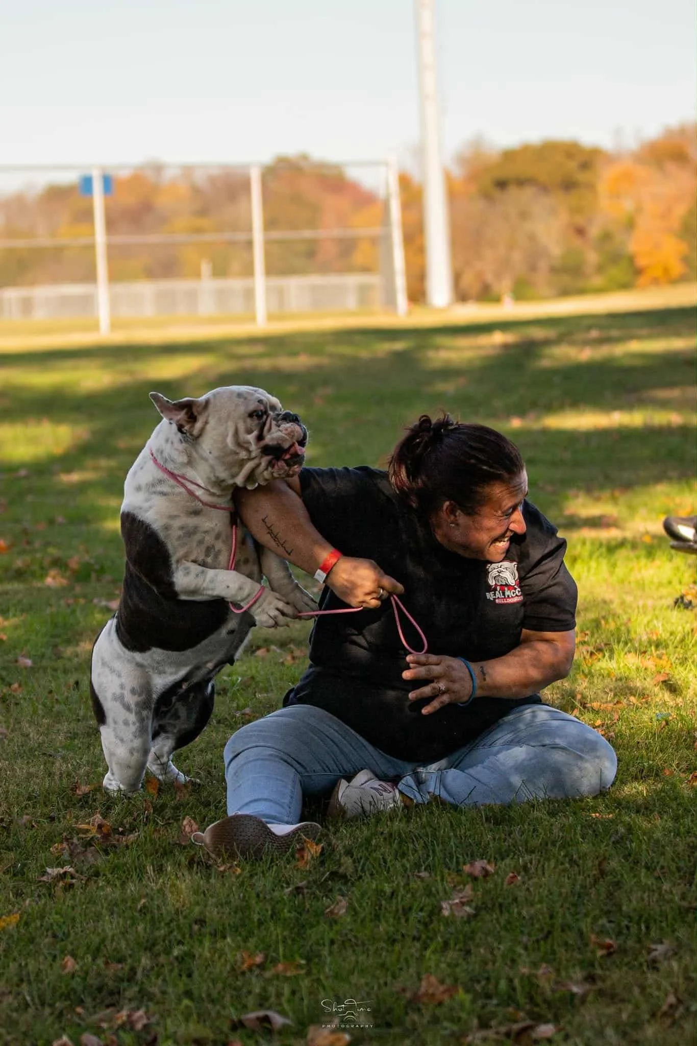 A man is sitting on grass outdoors with a bulldog jumping onto his back, both smiling and laughing. The background includes trees with fall foliage and a sports field with goal posts.