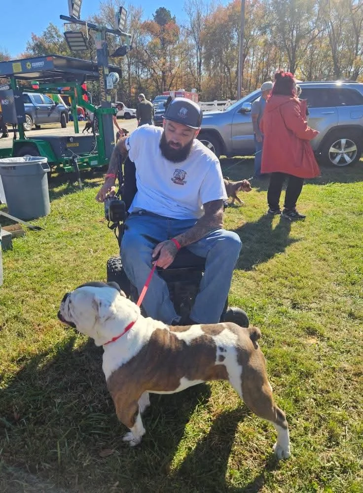 A bearded man in a white t-shirt and baseball cap sitting on a black chair outdoors, holding a leash attached to a brown and white bulldog. There are cars and people in the background, along with trees and a clear blue sky, suggesting a park or public event.